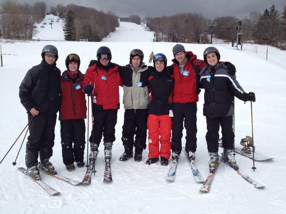 Fraternity brothers in ski gear standing together on a snowy slope, with ski lifts and trees in the background.