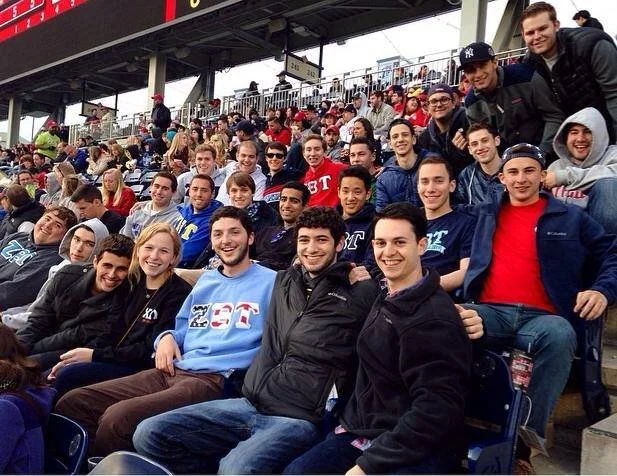 Fraternity brothers in stadium seating at an outdoor sporting event, smiling at the camera.
