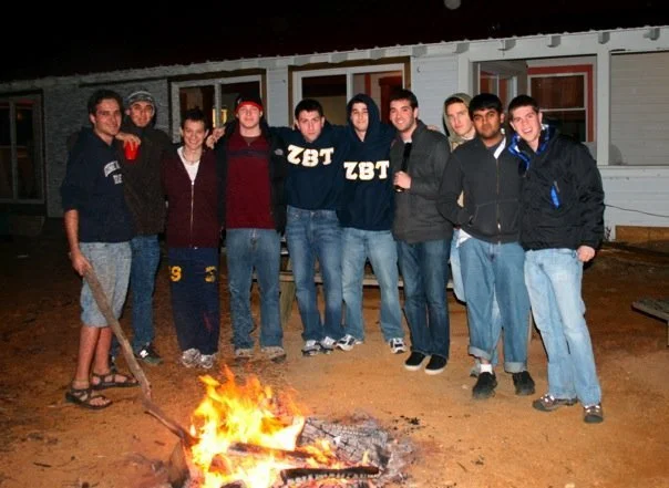 Fraternity brothers standing outdoors at night beside a campfire, with a building in the background.