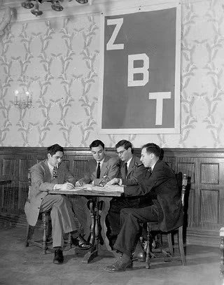 Fraternity founders sat at a table with ZBT letters in the background.