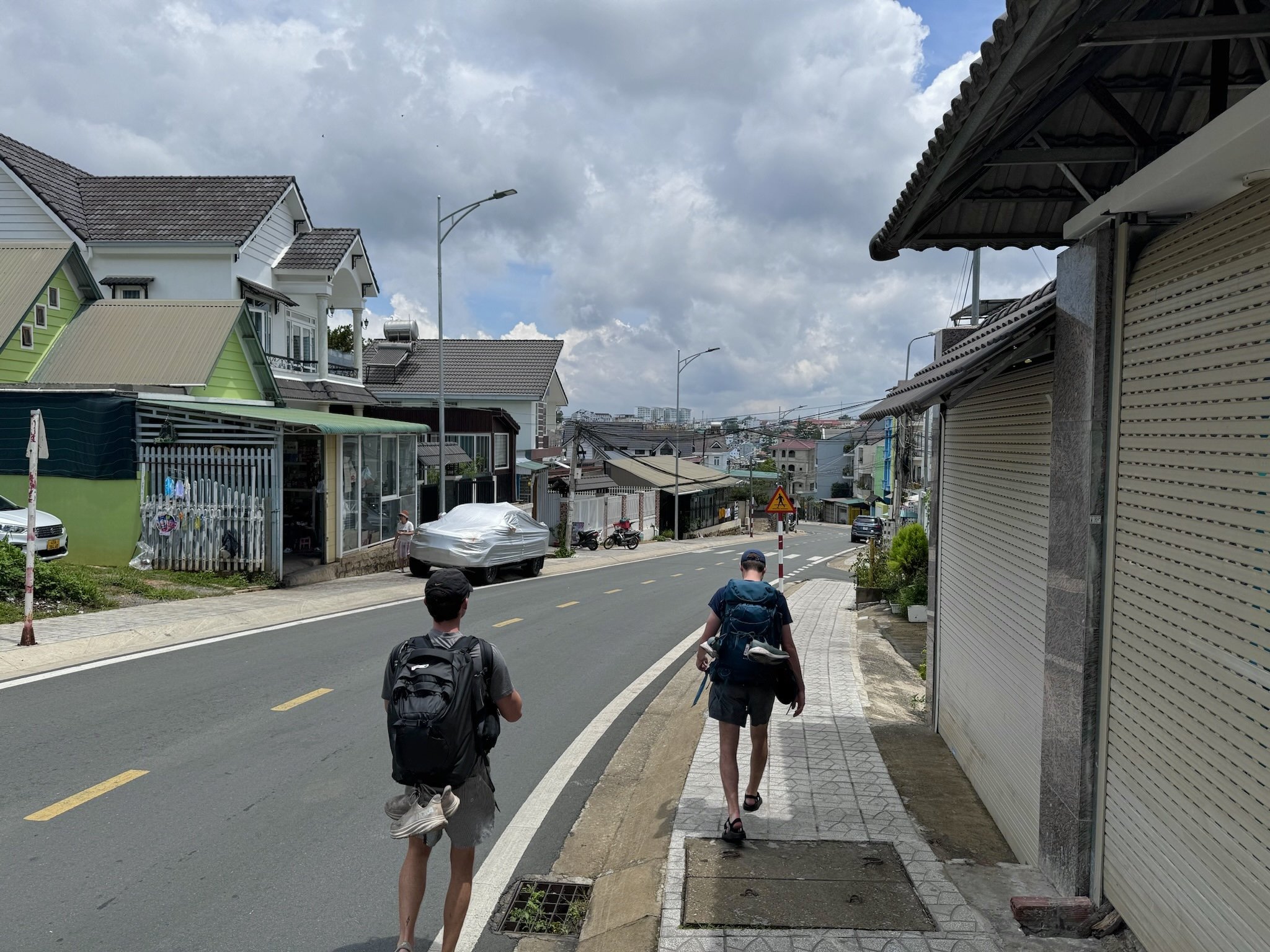 Two men with backpacks walking on a sidewalk along a hilly residential street, with houses on both sides and cloudy sky overhead.