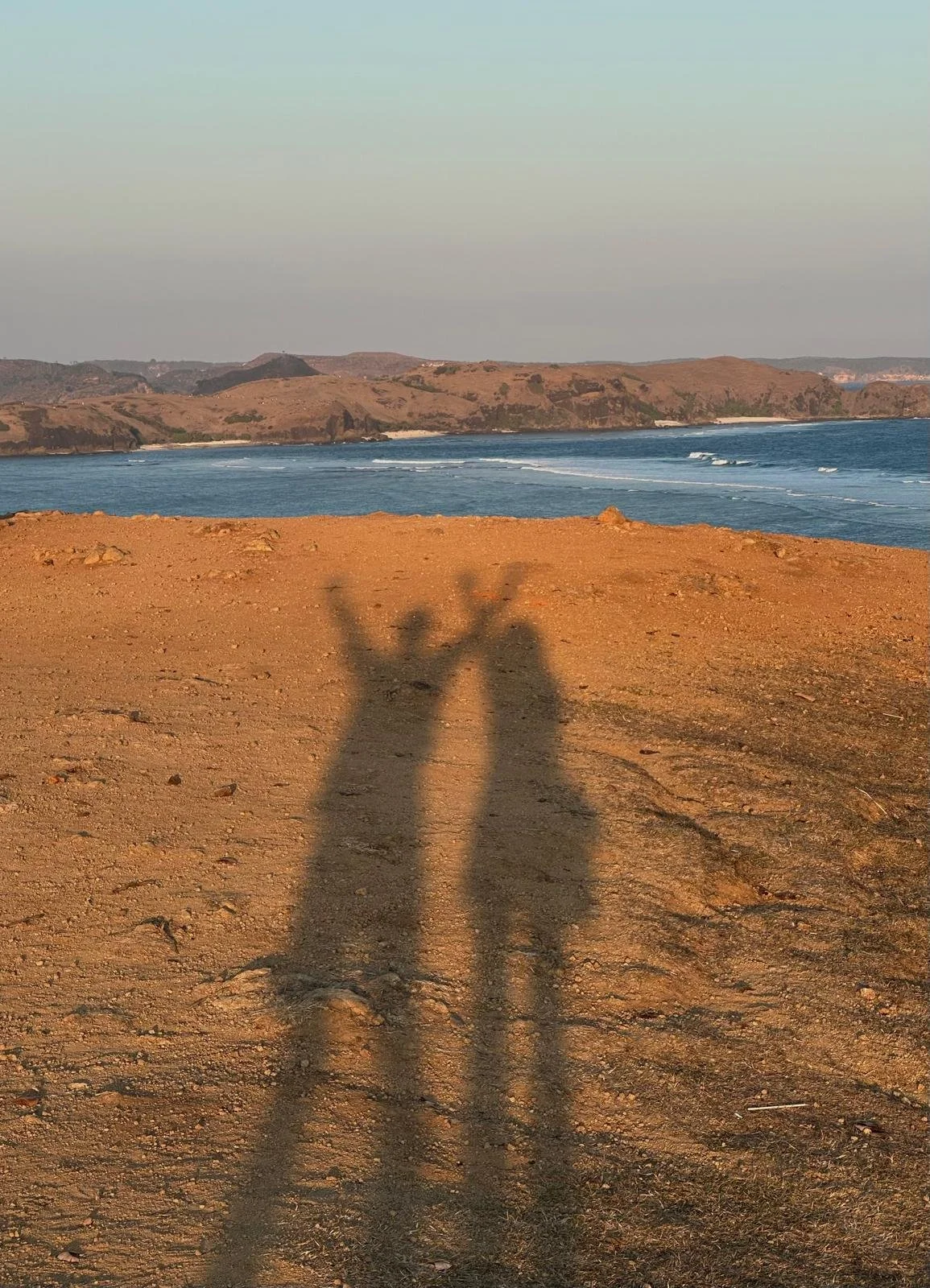 Shadows of two people making bunny ears and a peace sign on a dirt path at a beach with hills, water, and sky in the background.