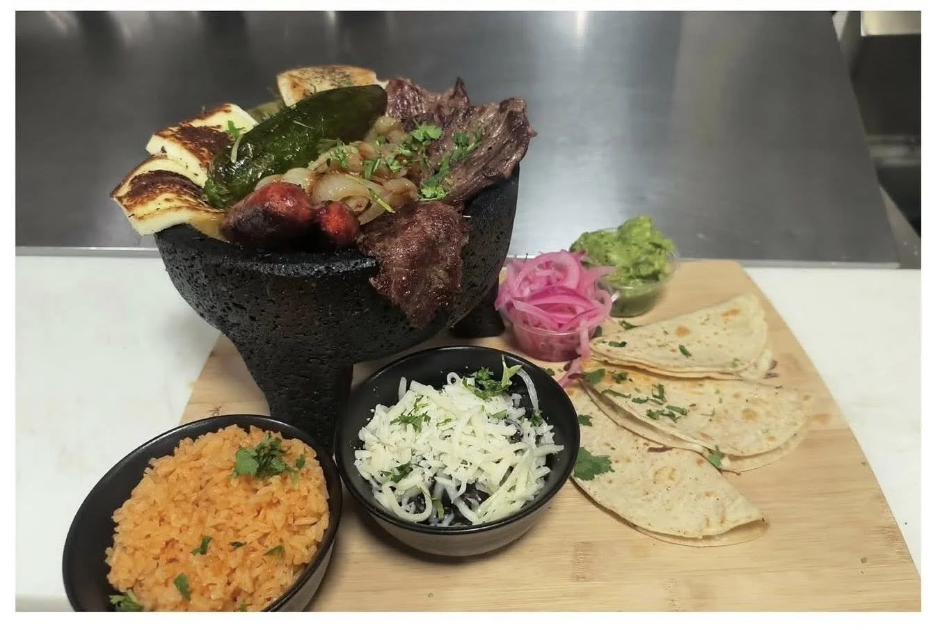 A serving of Mexican cuisine featuring a black molcajete filled with grilled meat, vegetables, and cheese, accompanied by rice, shredded cheese, pickled red onions, guacamole, and flour tortillas on a wooden board.