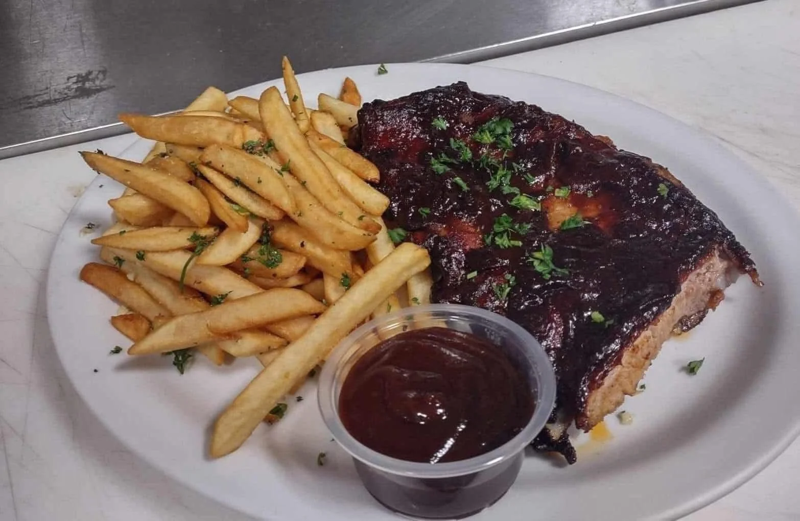 Pork ribs with barbecue sauce, seasoned French fries with herbs, and a side of barbecue sauce on a white plate.