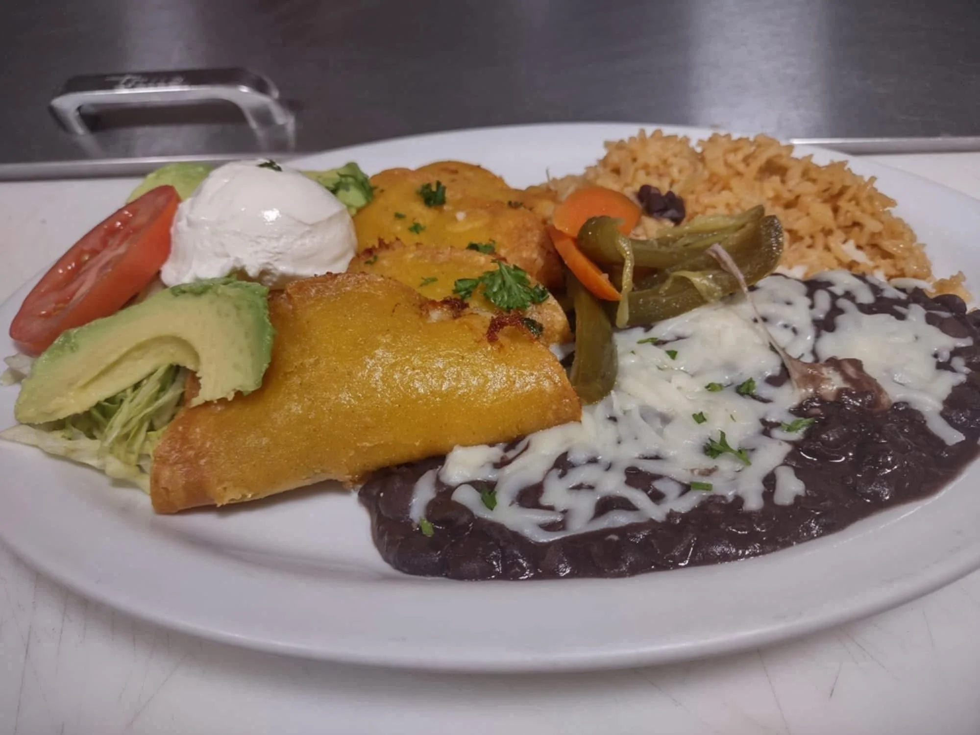 A plate of Mexican food with rice, black beans topped with melted cheese, beef or chicken enchilada with green sauce, fried plantain, sour cream, avocado slices, tomato and lettuce.