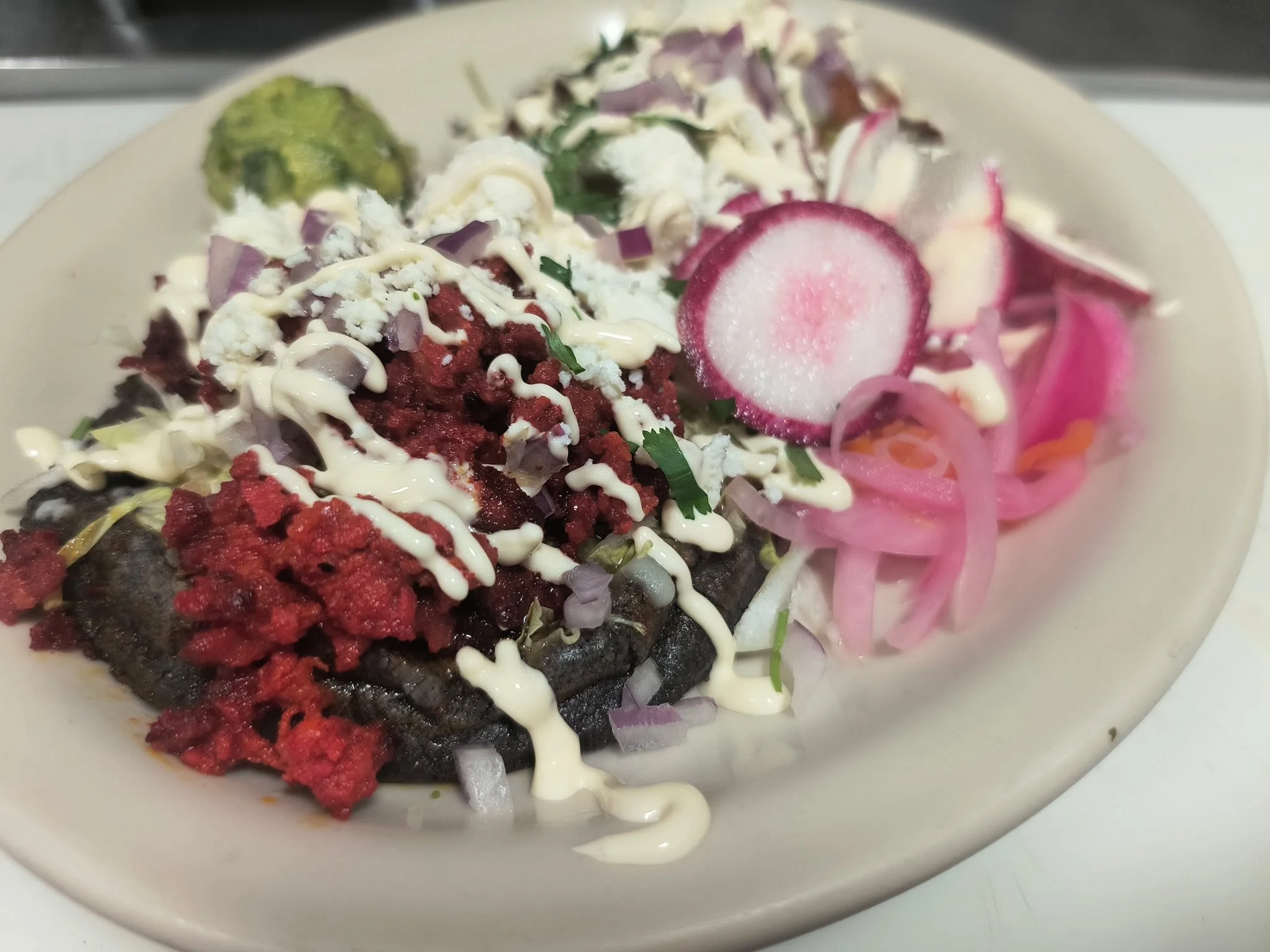 A plate of Mexican black bean tostada topped with red sauce, chopped onions, cilantro, sour cream, and thinly sliced radishes and pickled onions.