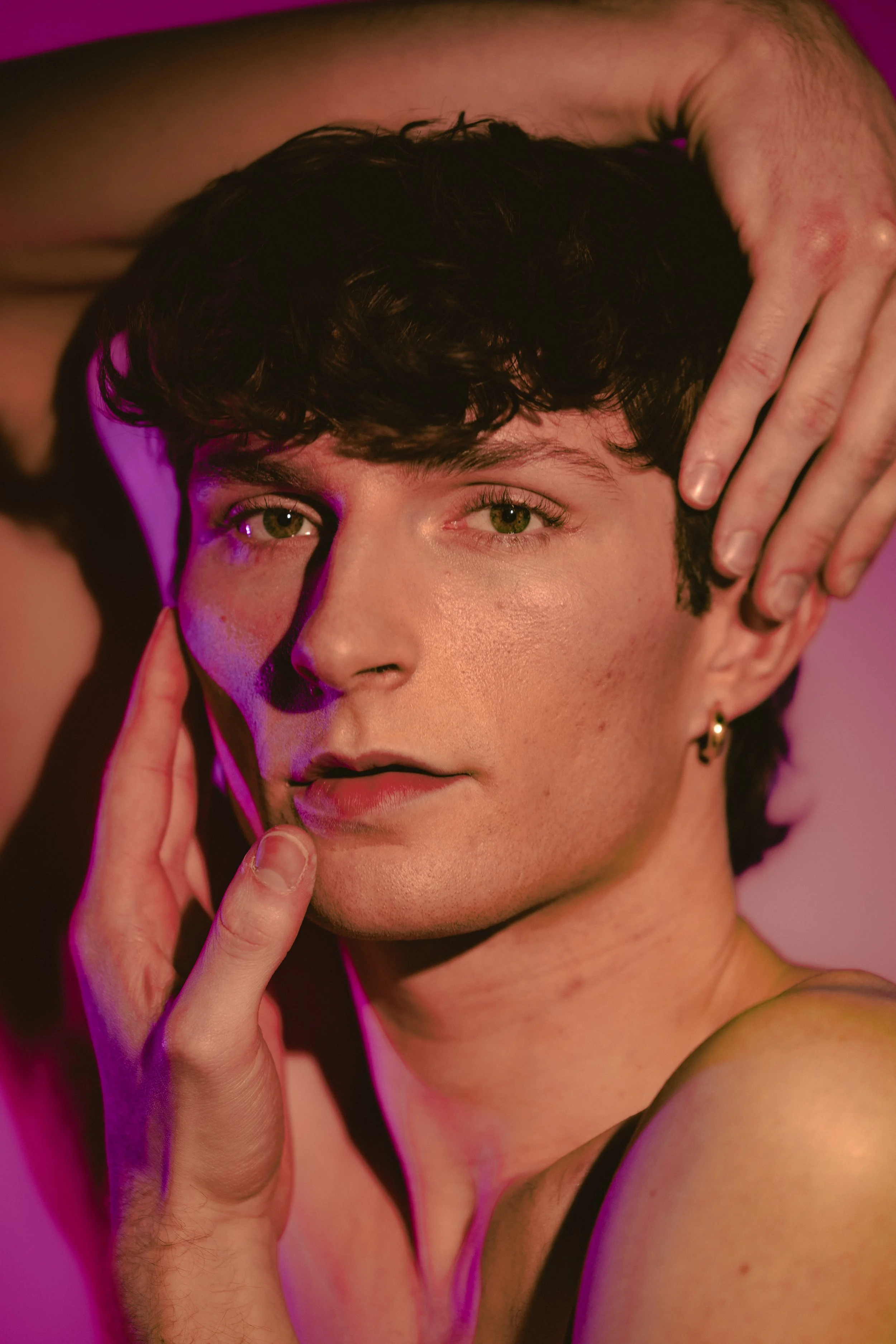 Close-up portrait of a young man with black curly hair, green eyes, and light skin, touching his face with fingers, pink and purple lighting.