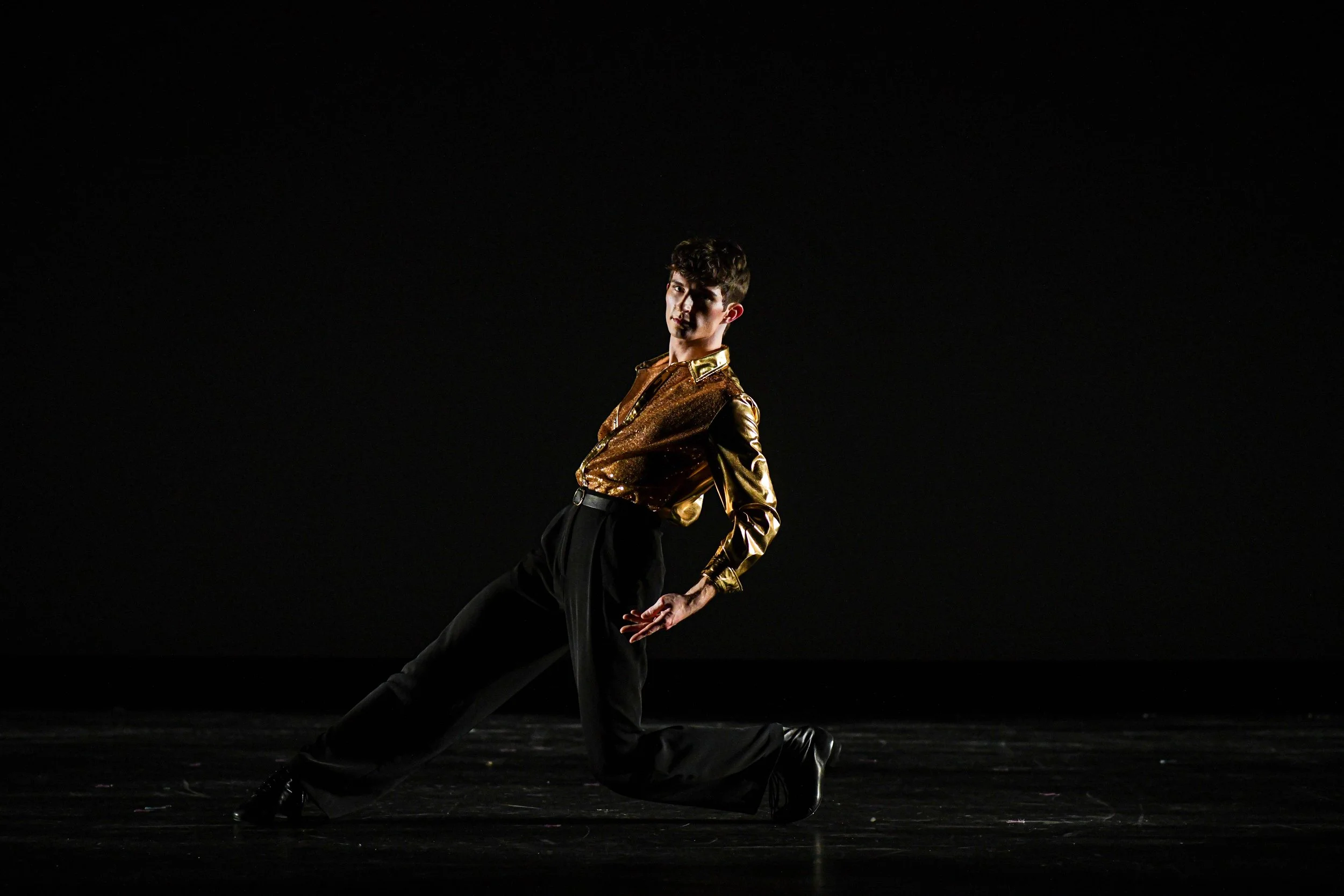 A young person performing a dance move on stage, wearing a shiny gold and brown top with black high-waisted pants, against a dark background.