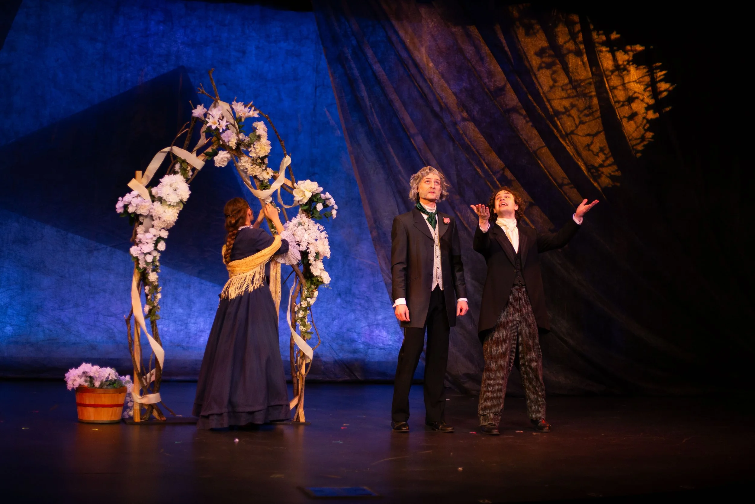 Three actors on stage during a theatrical performance, with two men dressed in period costumes and a woman adjusting a flower wreath on a wooden structure decorated with flowers and ribbons, set against a painted backdrop of trees.