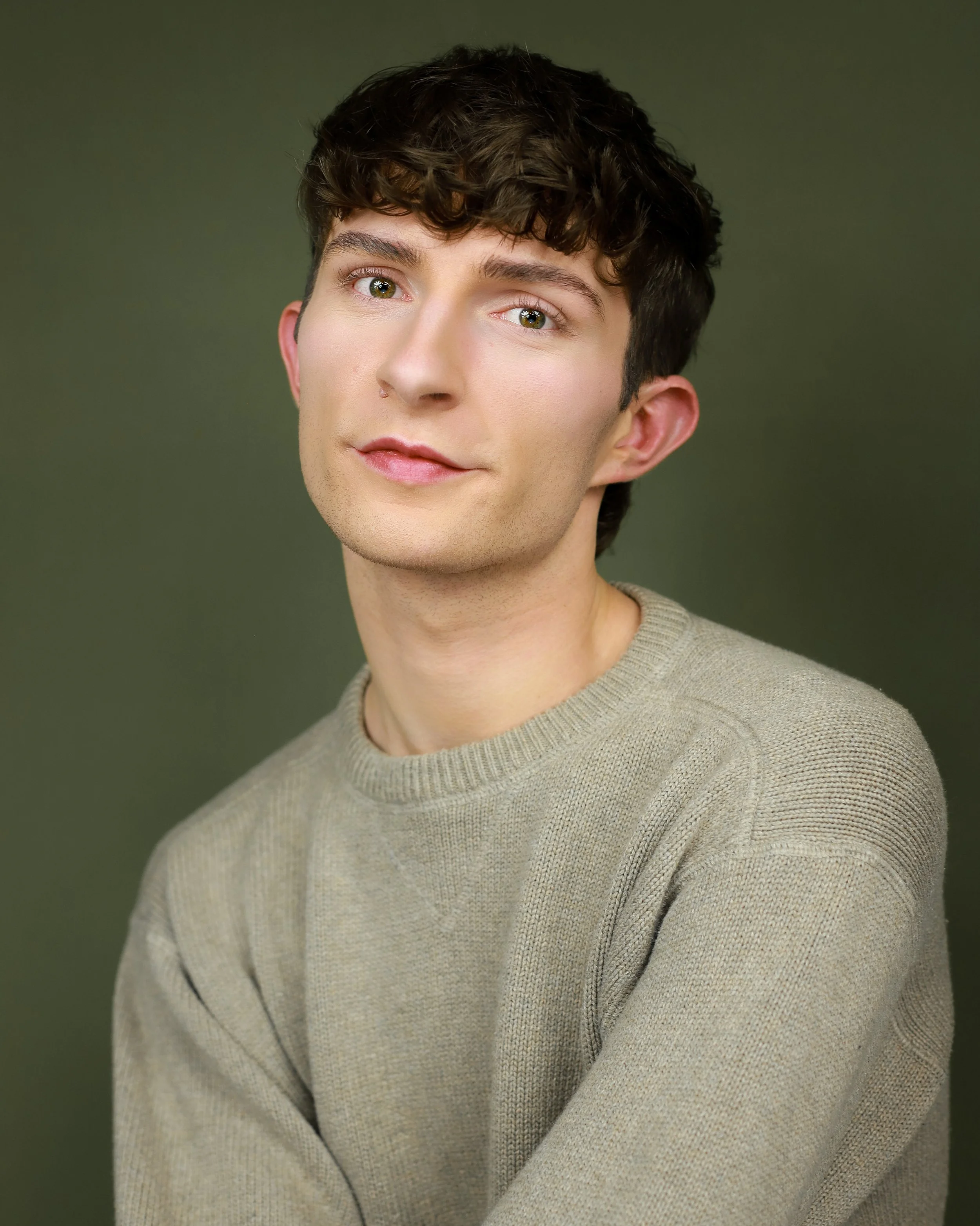 A young man with short curly dark hair and hazel eyes wearing a beige sweater, smiling softly against a plain green background.