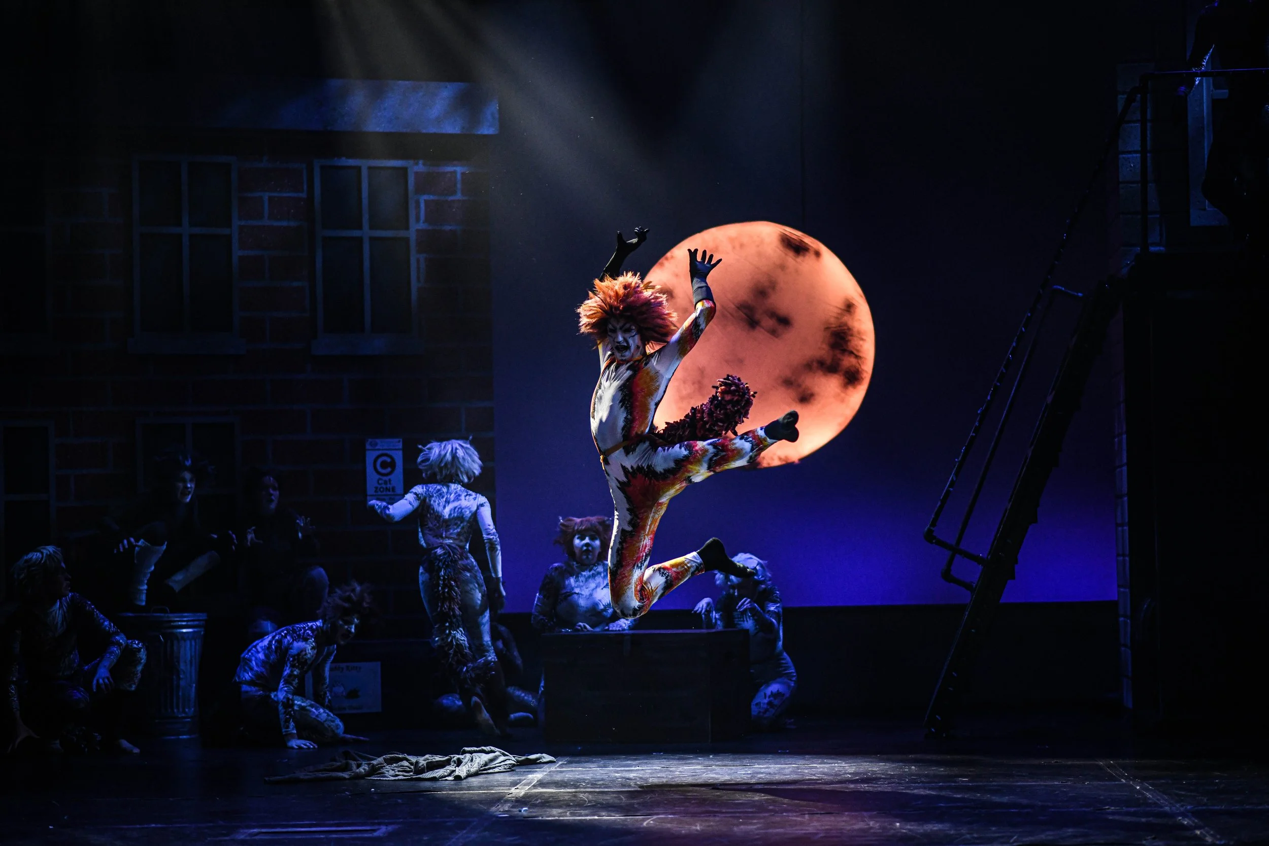 Performer with wild orange hair in colorful costume jumping in the air on stage with a large full moon backdrop, surrounded by other performers in animal costumes.