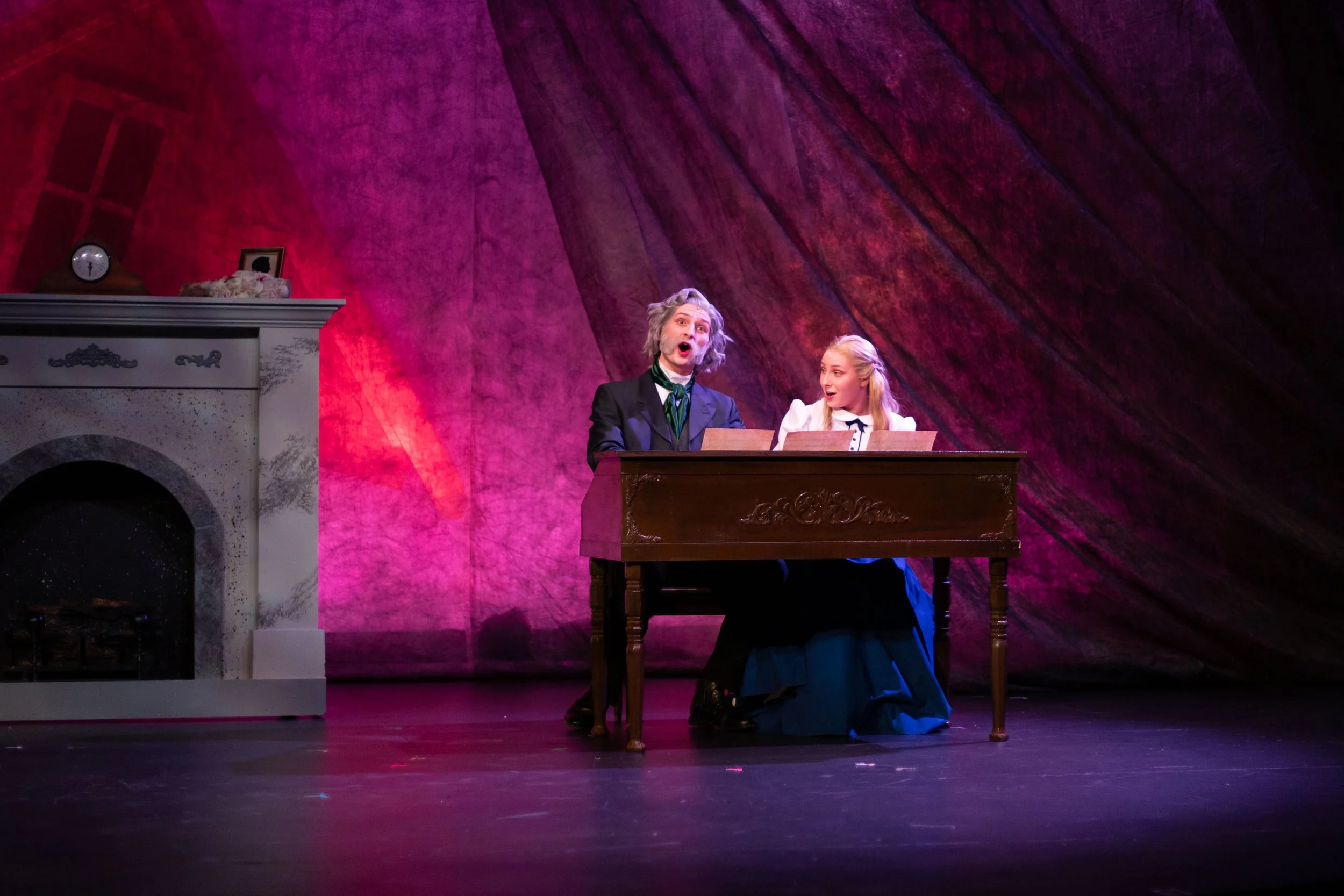 Two actors in period costume performing on stage, one with gray hair and a surprised expression, the other with blonde hair, seated at a desk, in a theatrical play setting with a pinkish-purple backdrop.