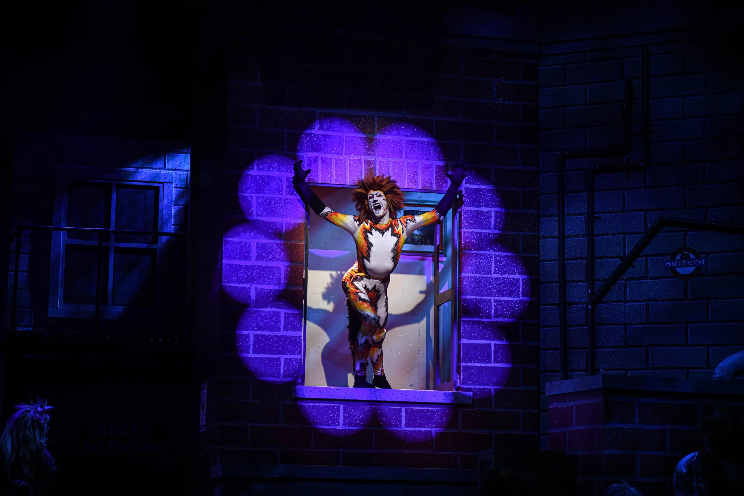A performer dressed in a colorful, tiger-striped costume and face paint, standing on a stage with arms extended, illuminated by purple and blue spotlights against a dark brick wall background.