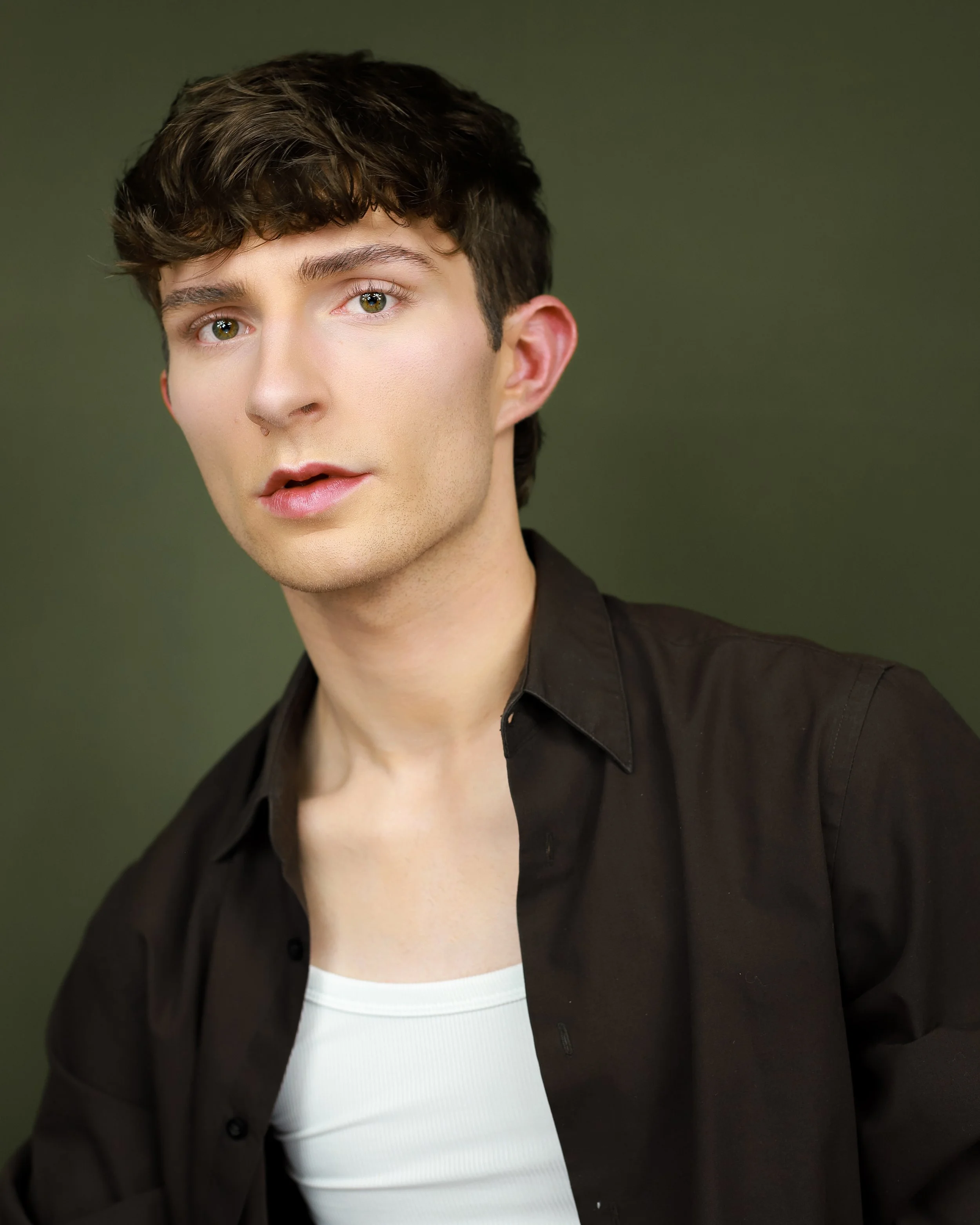 A young man with brown hair and hazel eyes wearing a black shirt over a white T-shirt with a green background.
