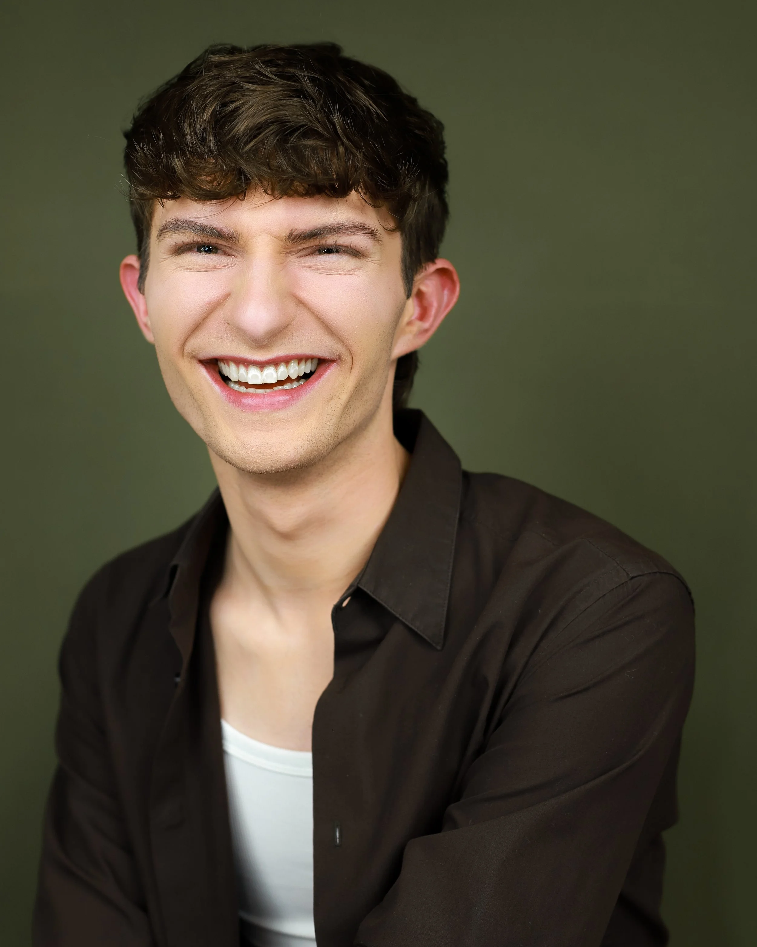 A young man with dark brown, curly hair, fair skin, and blue eyes, smiling and showing teeth, wearing a white shirt under a black jacket, against a plain green background.