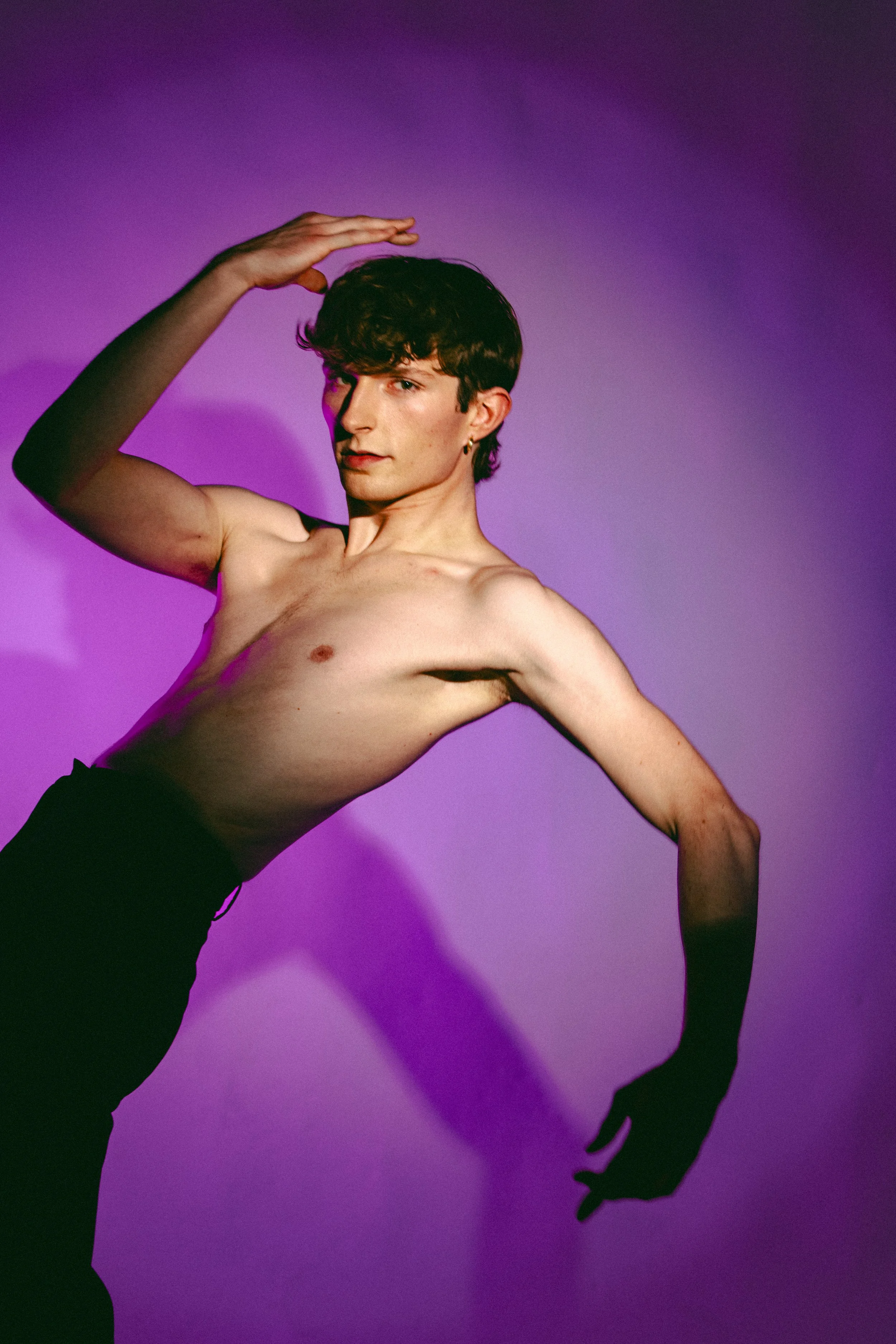 A shirtless young man with dark, short, curly hair and light skin poses against a purple background, with his right arm raised above his head and his left arm bent, touching his forehead.