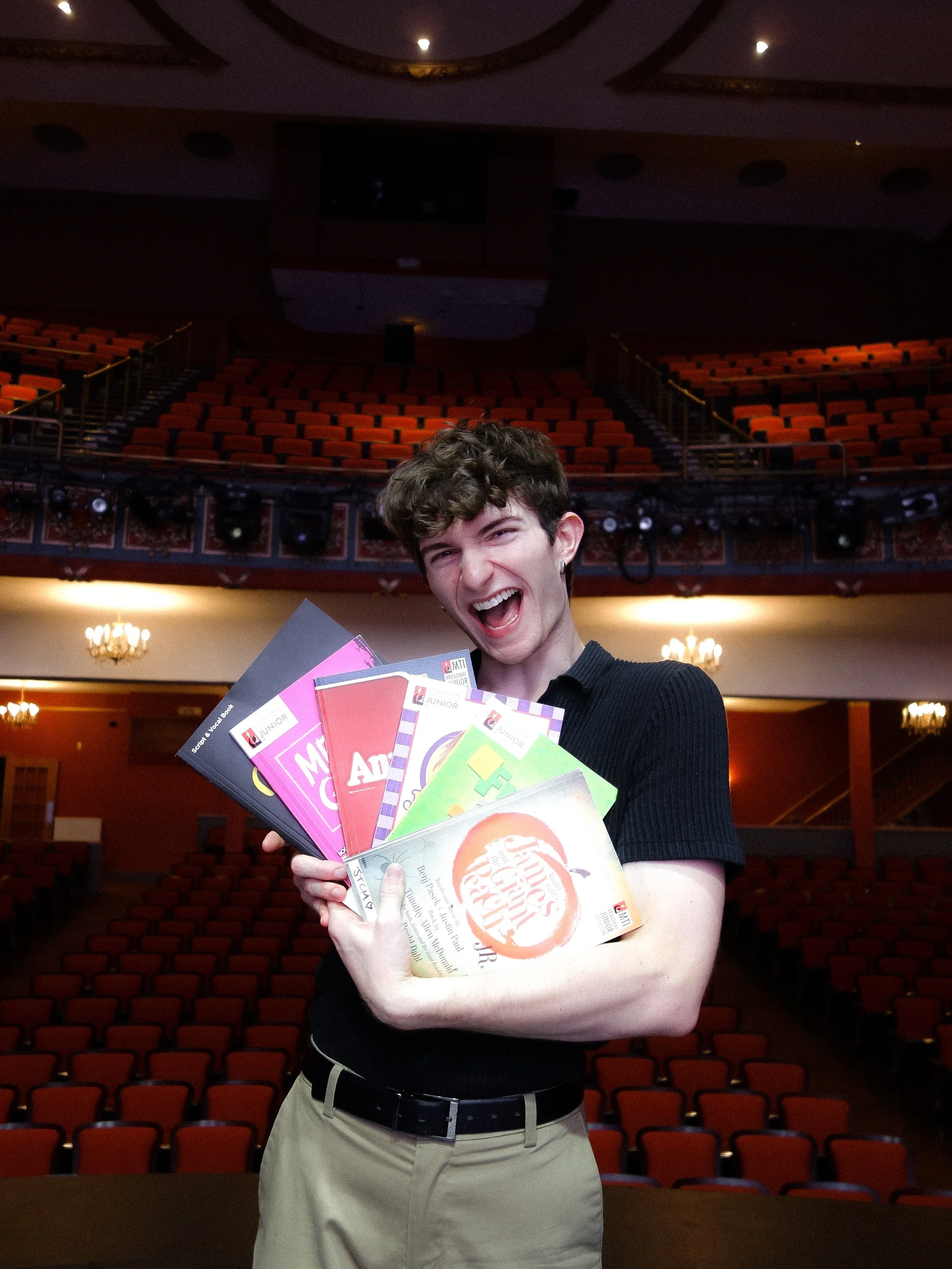 A young man joyfully holding colorful booklets in a large auditorium with empty red seats and ornate chandeliers in the background.