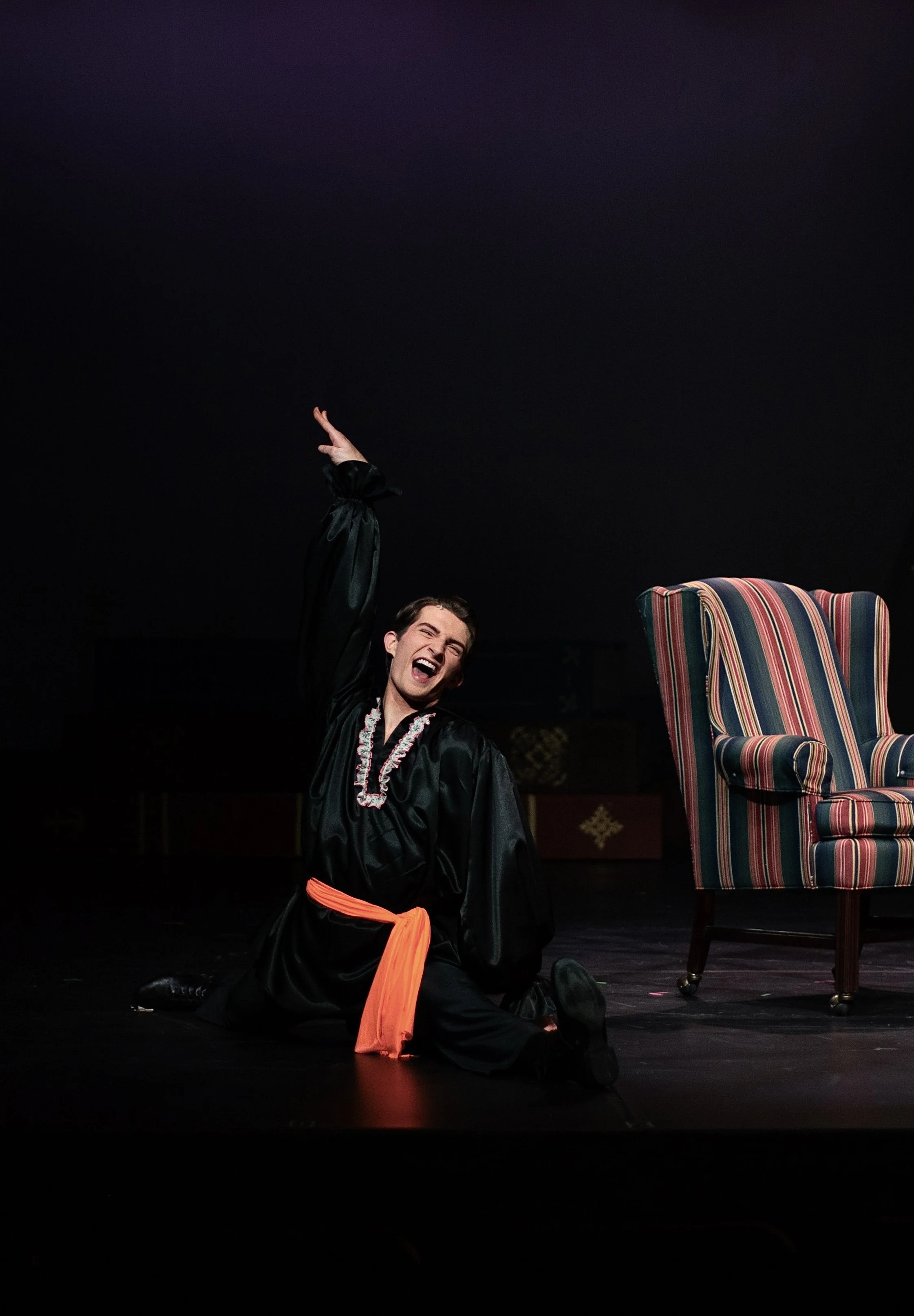 Person in a black graduation gown with an orange sash kneeling on stage, smiling and gesturing upward with one hand, with a striped armchair beside them on a dark stage.