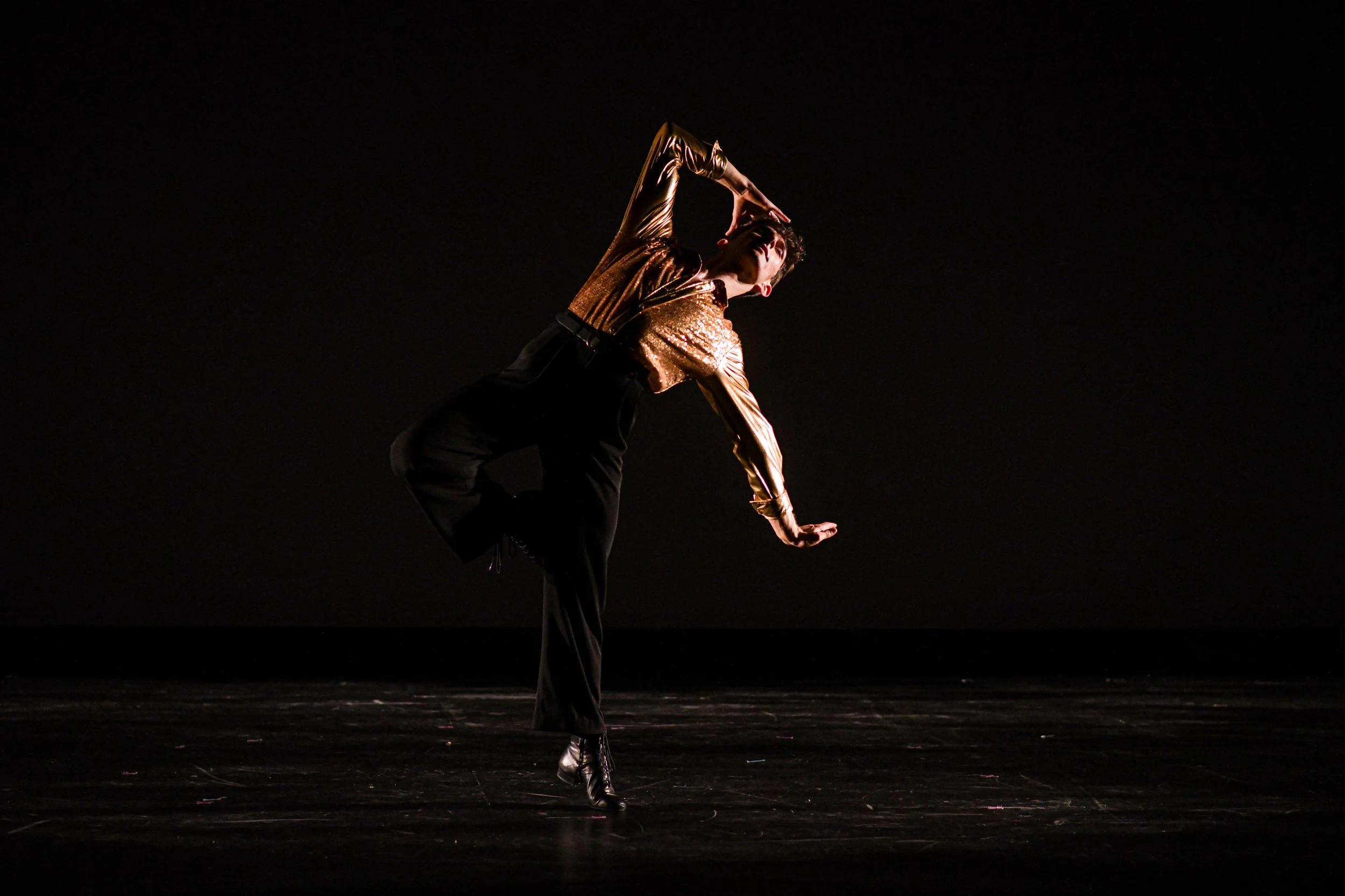 A performer in a shiny gold and black costume dances on stage against a black background, striking a pose with one leg lifted and one arm bent over the head.