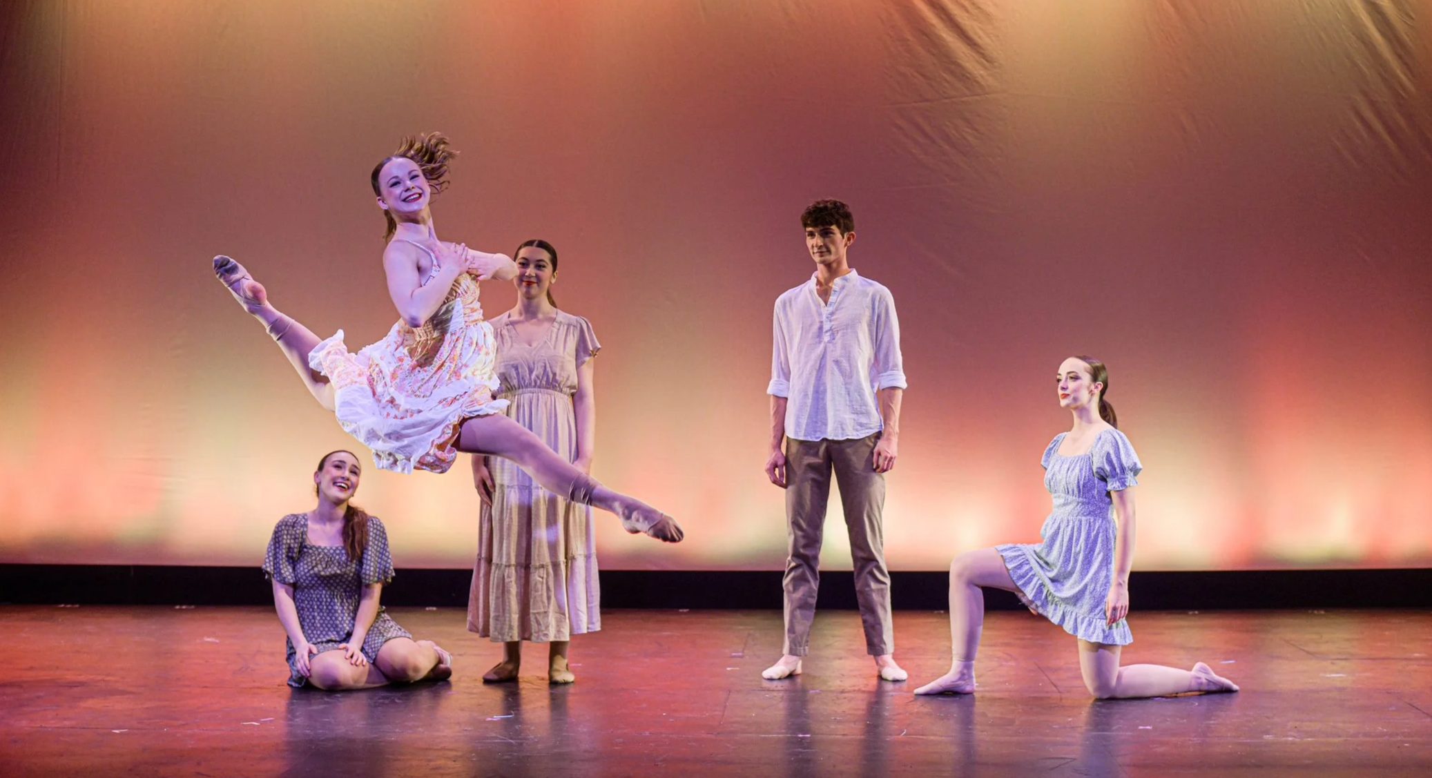 Ballet dancers on stage, with one dancer mid-leap, surrounded by four other dancers, against a warm, gradient backdrop.