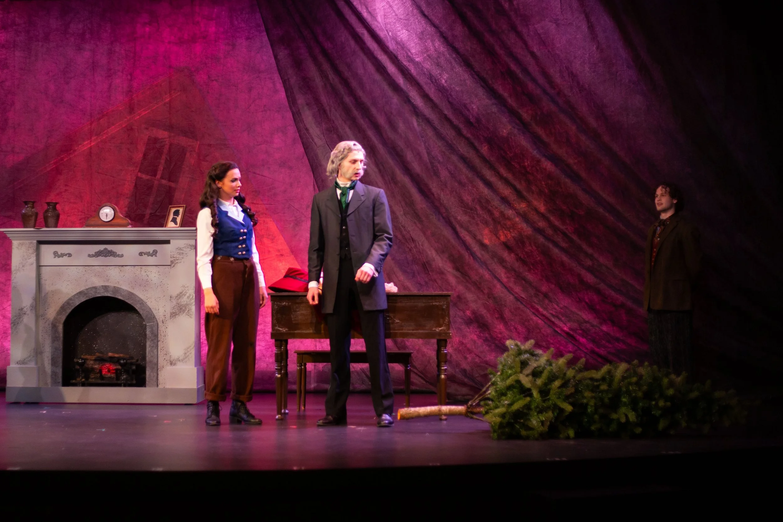 Three actors on stage performing a scene in front of a pink backdrop with a fireplace, a mantle with vases, a clock, and framed photo, and a bunch of green foliage at the front of the stage.