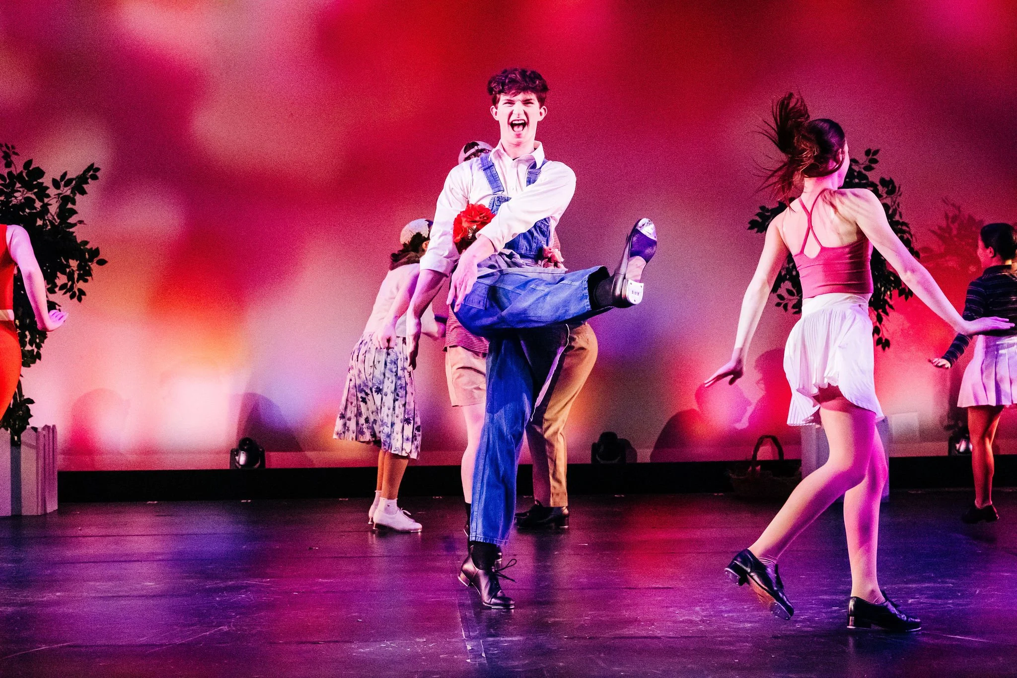 People performing a dance on stage with colorful lighting, some dancers in mid-movement, and a red and purple background.