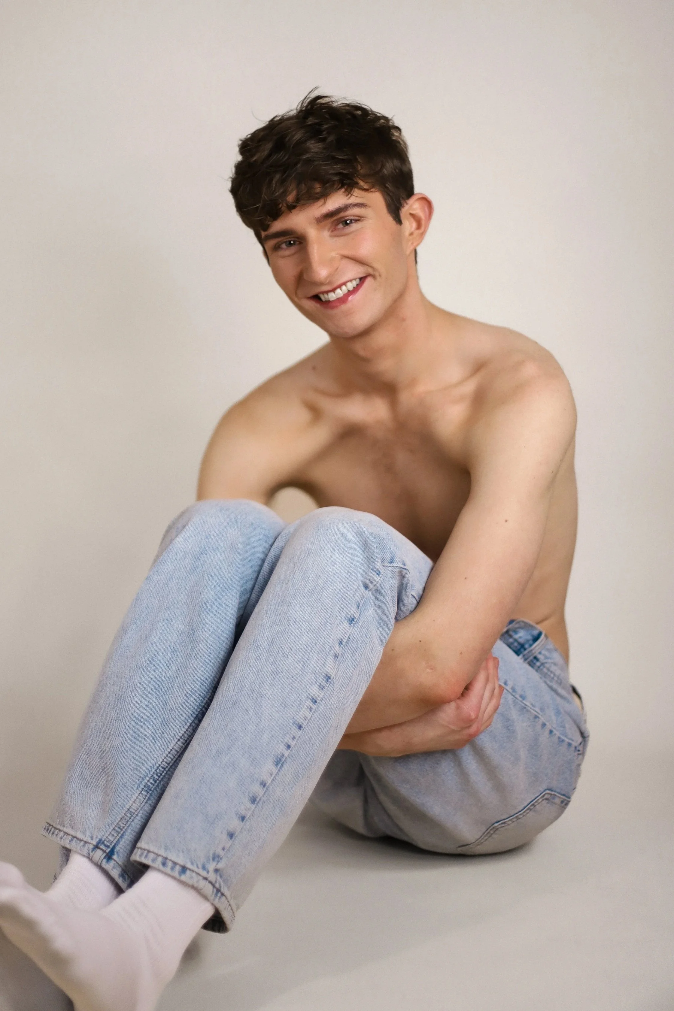 A young man with short dark curly hair and fair skin, smiling and sitting with knees pulled up, wearing light blue jeans and white socks, shirtless, against a plain beige background.