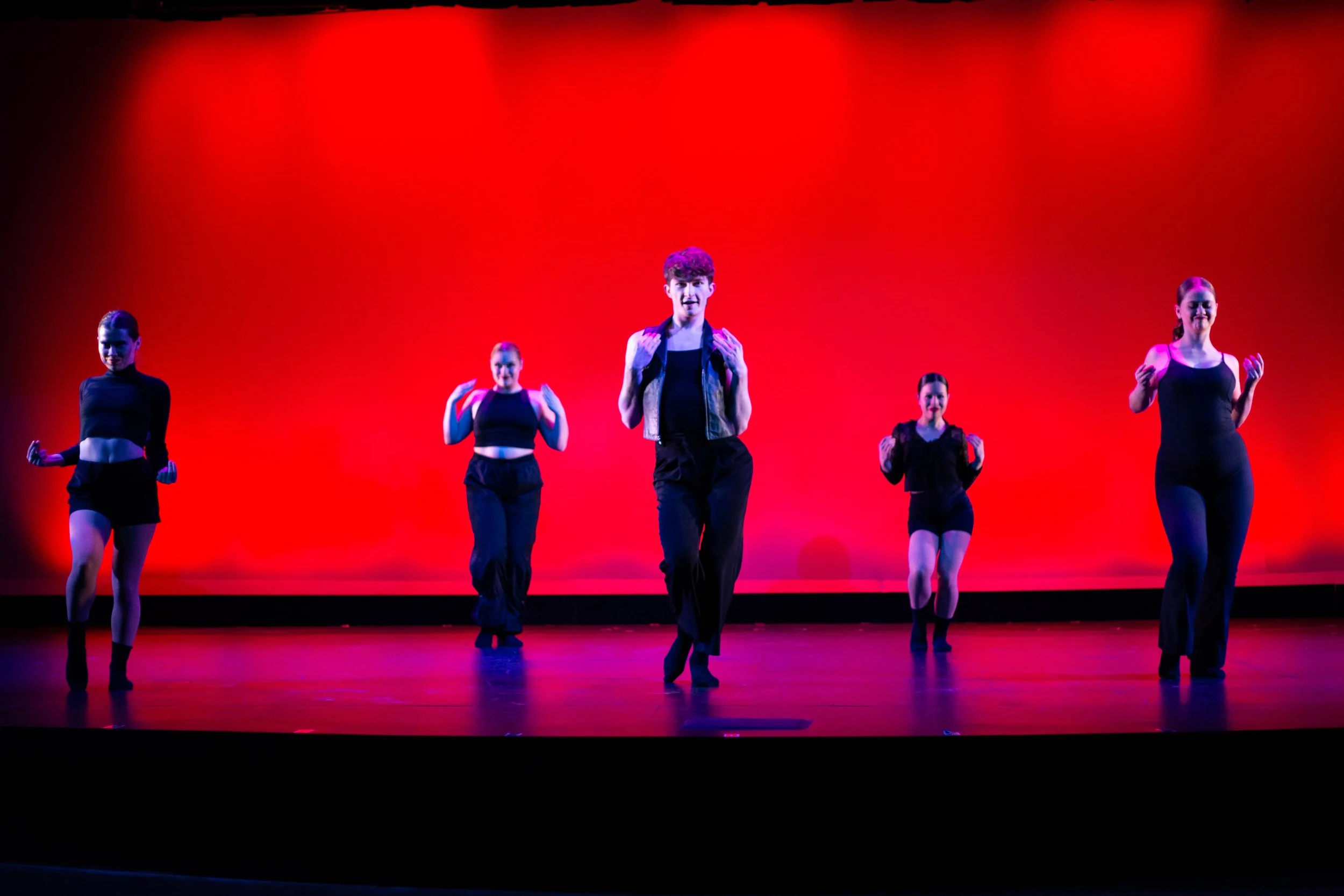 Five dancers on stage with a red background, mid-performance, wearing black clothing. The dancers are arranged with one in the center and two on each side, all smiling or with focused expressions.