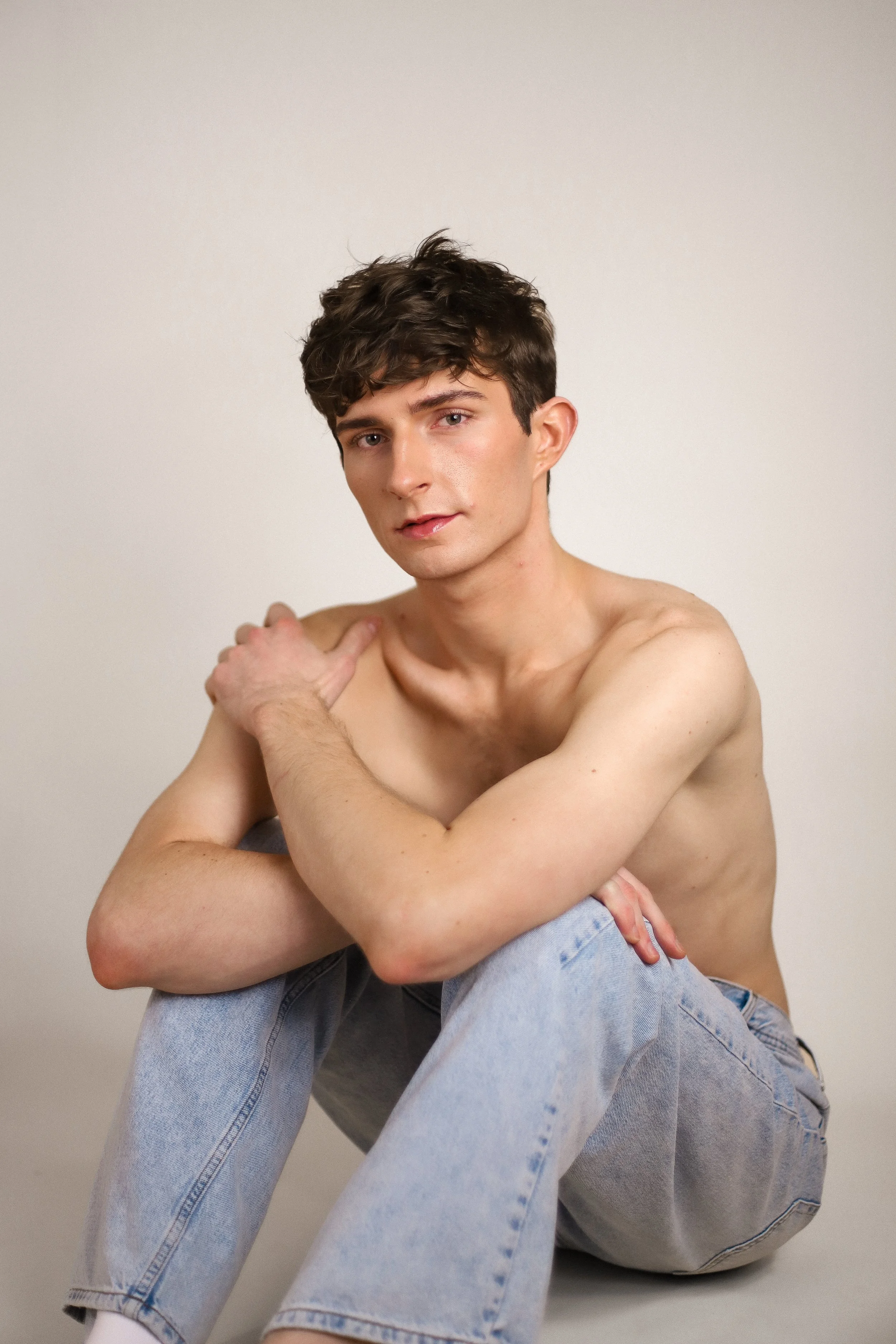 A young man with short, tousled brown hair, fair skin, and blue eyes is sitting against a plain light grey background. He is shirtless, wearing light blue jeans, and has a gentle, relaxed expression. He is resting his left arm on his knee and touching his right shoulder with his right hand.