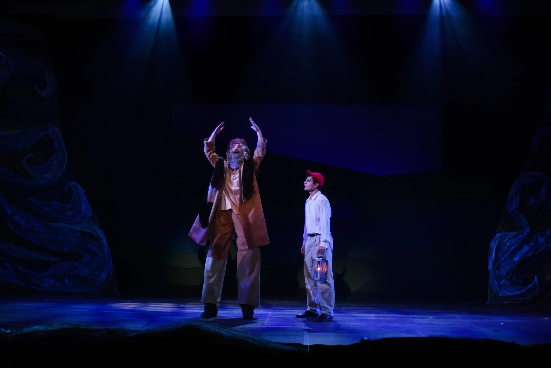 Two actors perform on stage with dark background, one with a long beard and traditional clothing gesturing upward, the other with glasses, a red cap, and a lantern watching attentively.