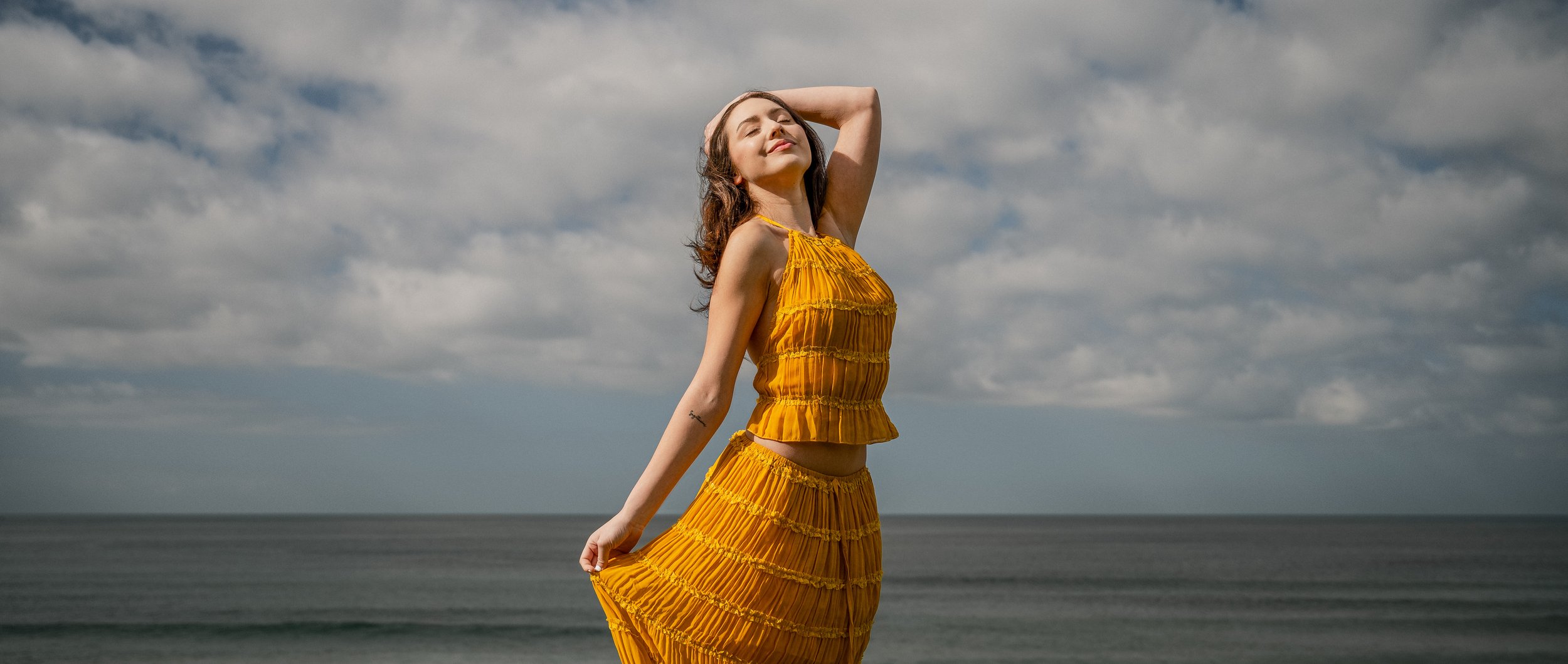 A woman in a yellow dress standing on the beach with her eyes closed and one arm raised behind her head, with a cloudy sky and ocean in the background.