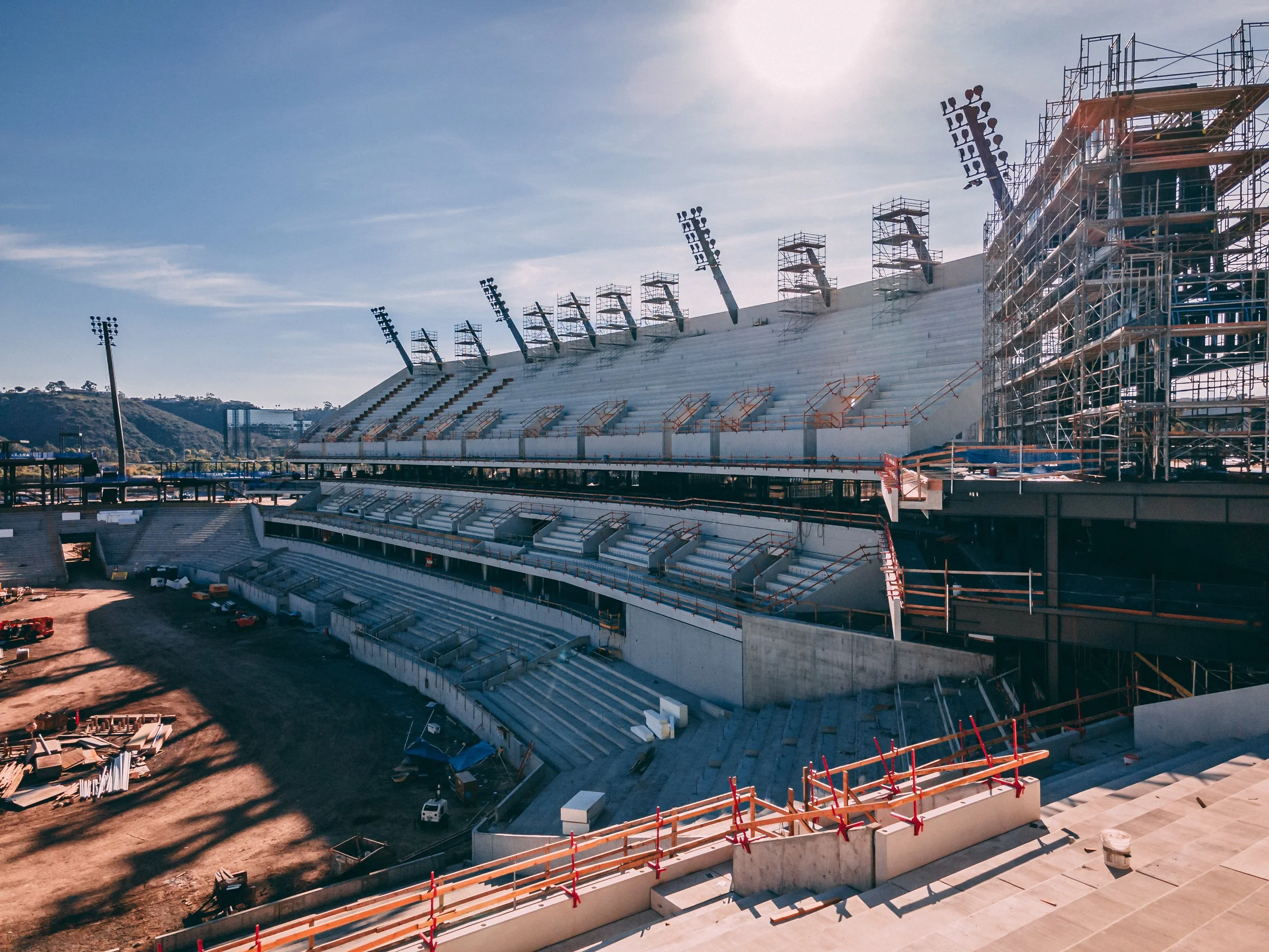 Under construction Snapdragon Stadium with seating and scaffolding, floodlights, and blue sky.