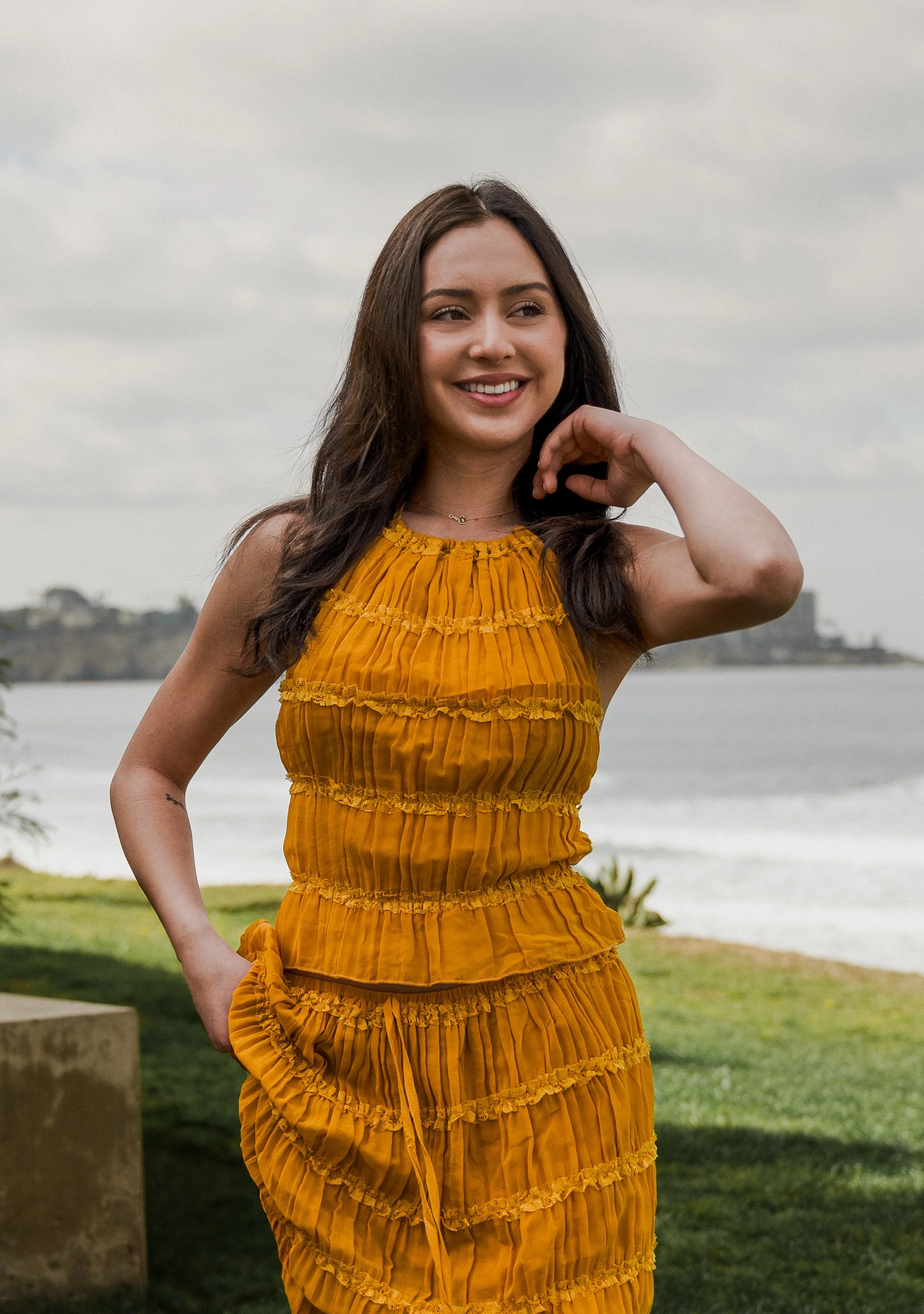 A young woman with long dark hair in a yellow dress with ruffles, smiling outdoors near a body of water on a cloudy day.