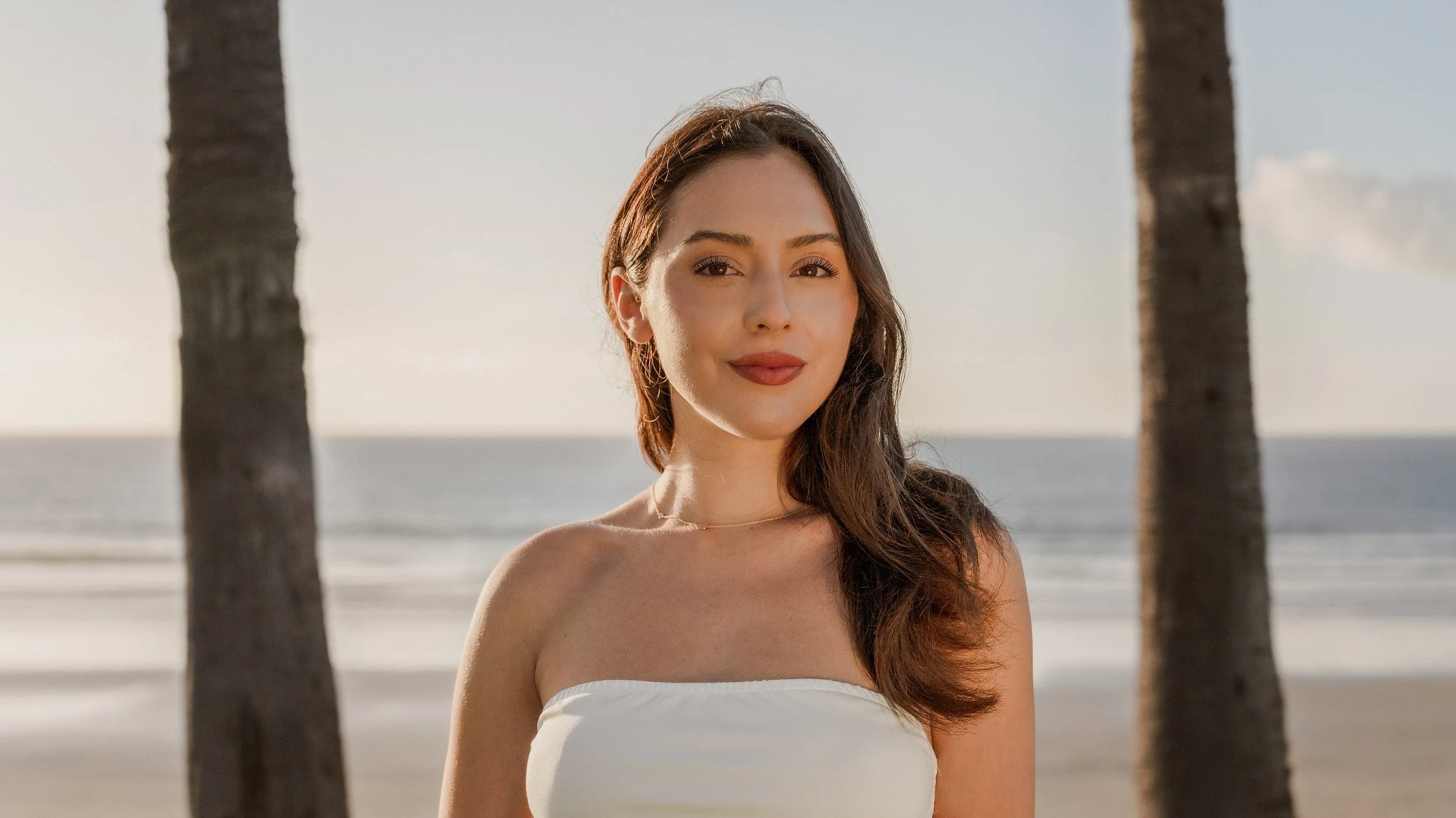 A woman standing on the beach between two palm trees during sunset, wearing a white strapless top and smiling softly.