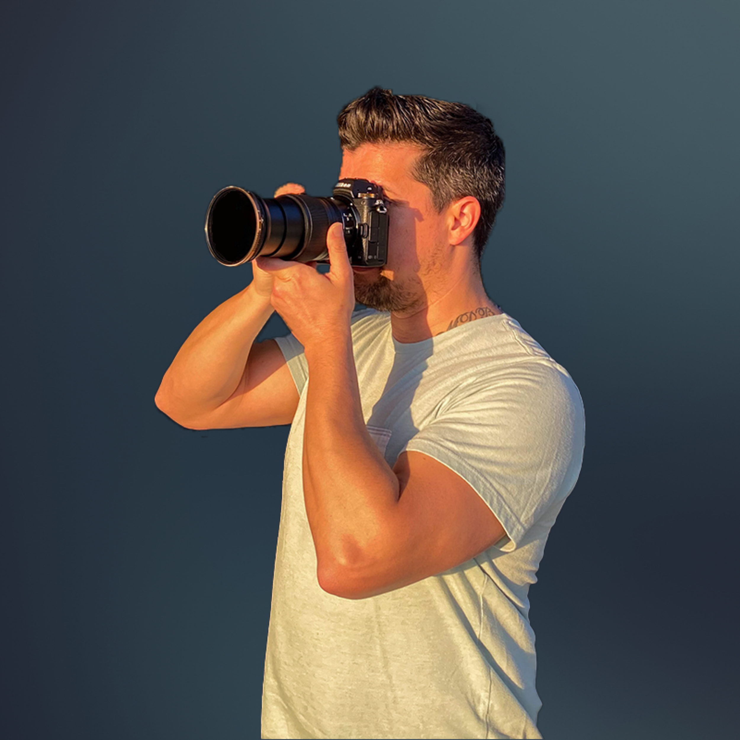 A man with short dark hair, wearing a light-colored t-shirt, is taking a photo with a professional camera against a dark background.