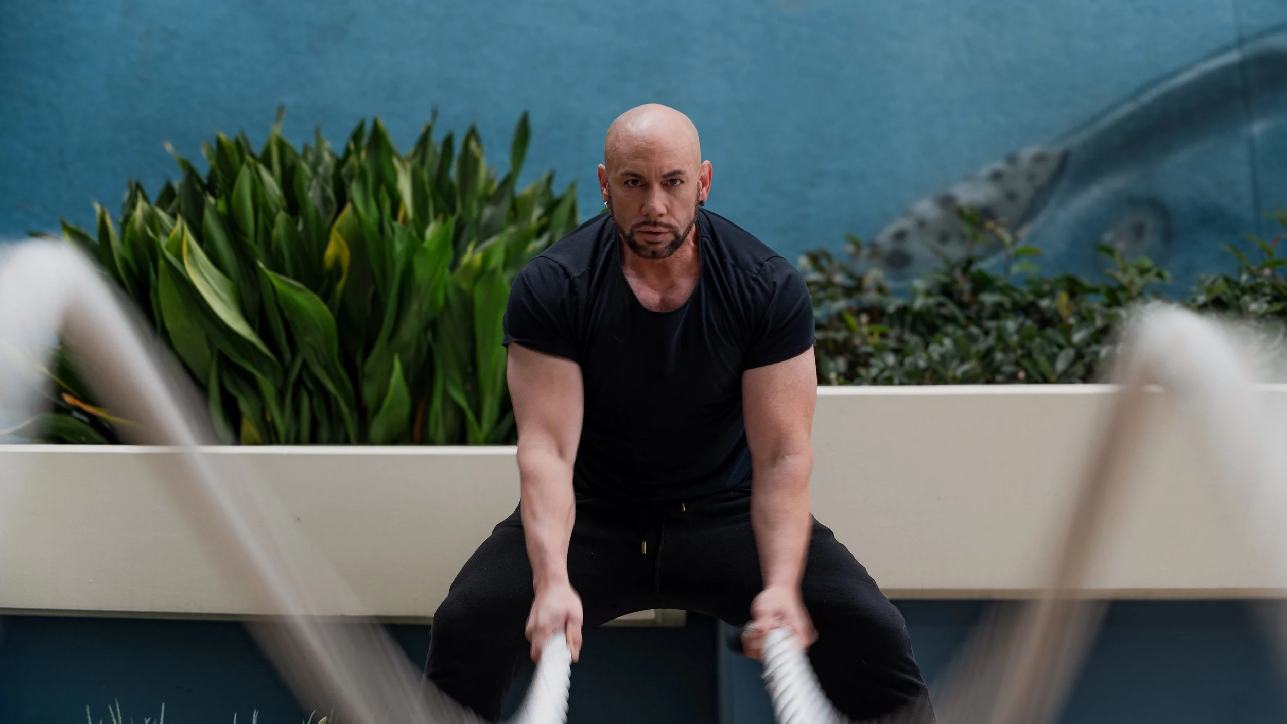 A muscular man with a bald head and beard, wearing a black t-shirt and black pants, exercises with battle ropes in an outdoor setting with green plants and a blue wall background.
