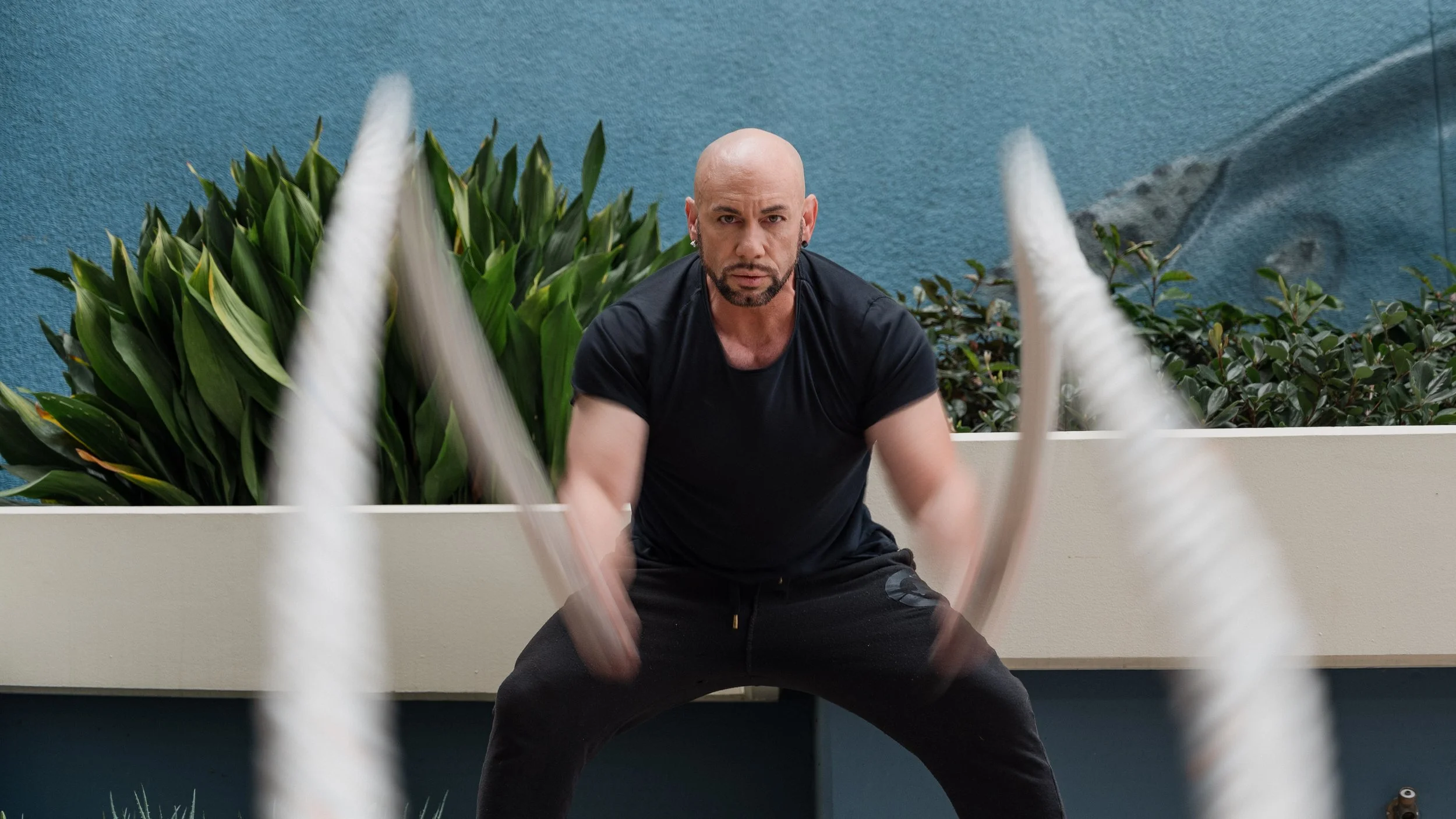 A man with a bald head, beard, and earrings wearing a black t-shirt and black pants, exercising with battle ropes in an outdoor area with green plants and a textured blue wall in the background.