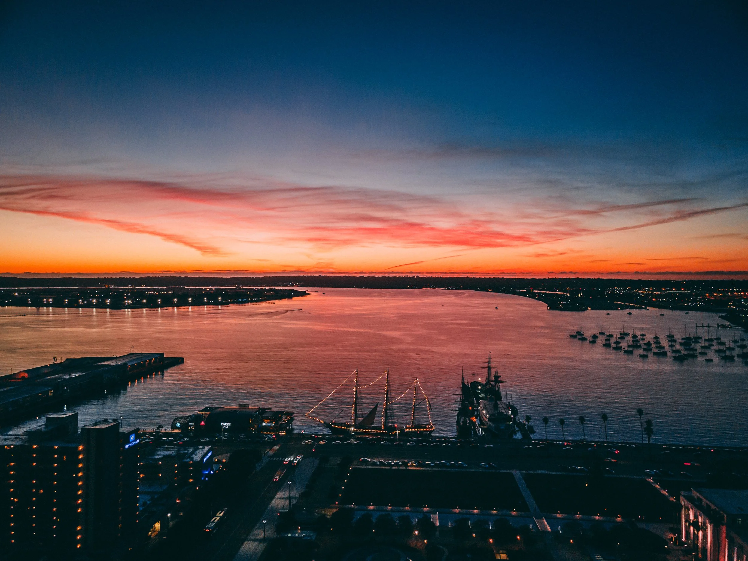 Sunset over a harbor with boats and yachts, city buildings, and a cloudy sky with orange, pink, and blue hues at the San Diego Harbor.
