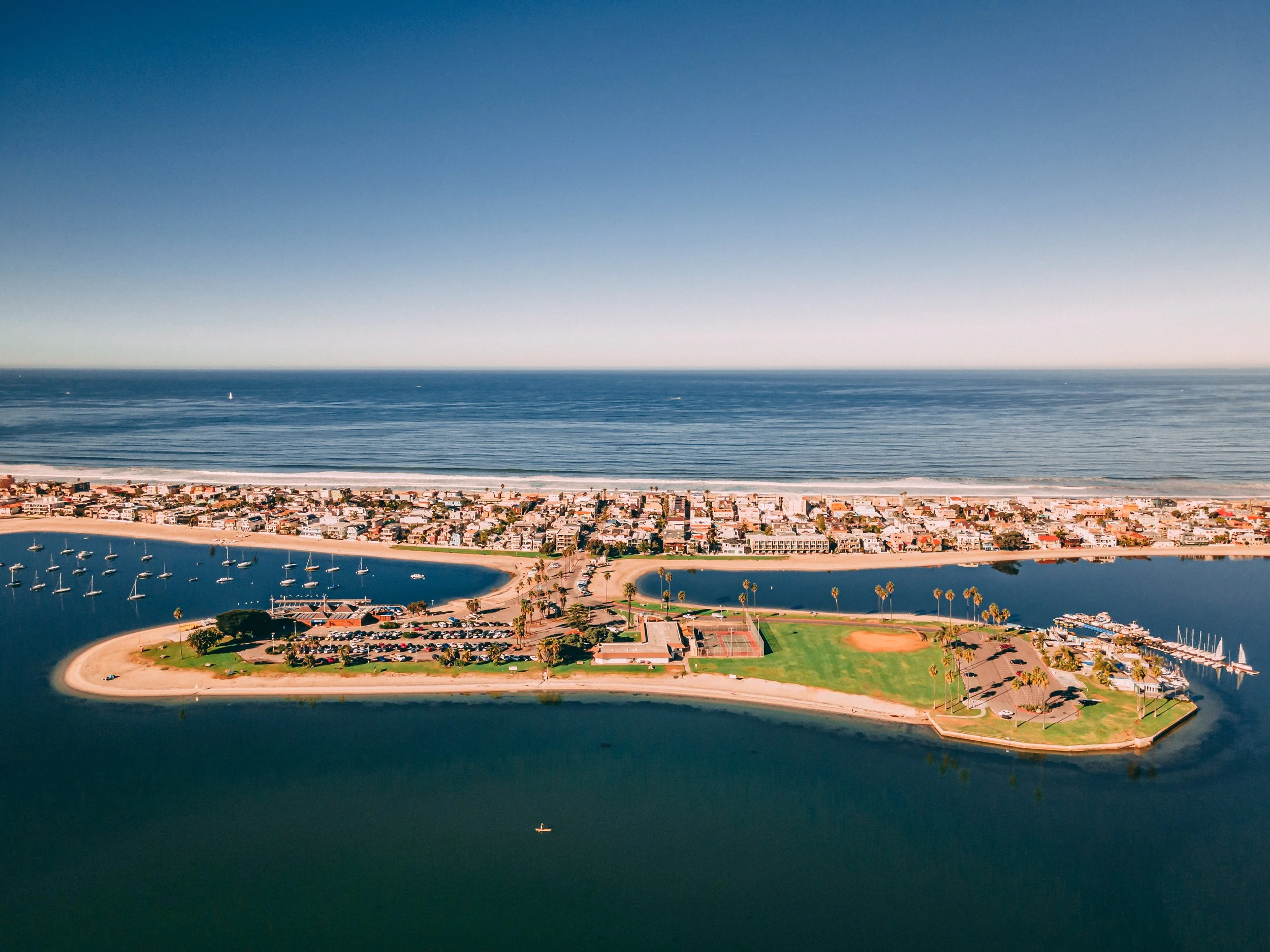 Aerial view of a coastal city with a peninsula featuring a park, marina, and baseball field, surrounded by a lagoon and ocean, with a dense residential area in the background.