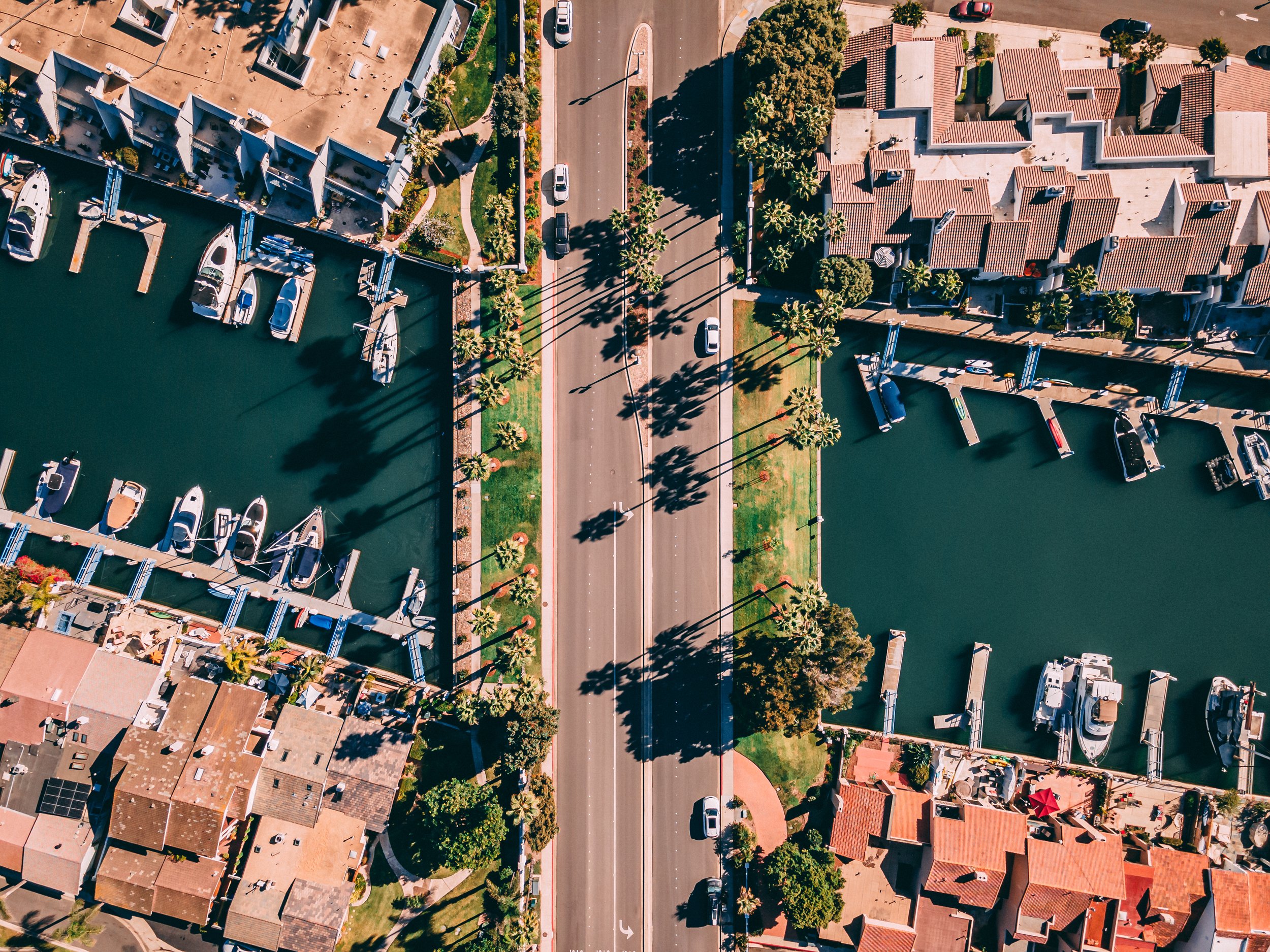 Aerial view of a marina in the Coronado Cays with boats docked along the water, a road with cars, and residential buildings with red-tiled roofs.