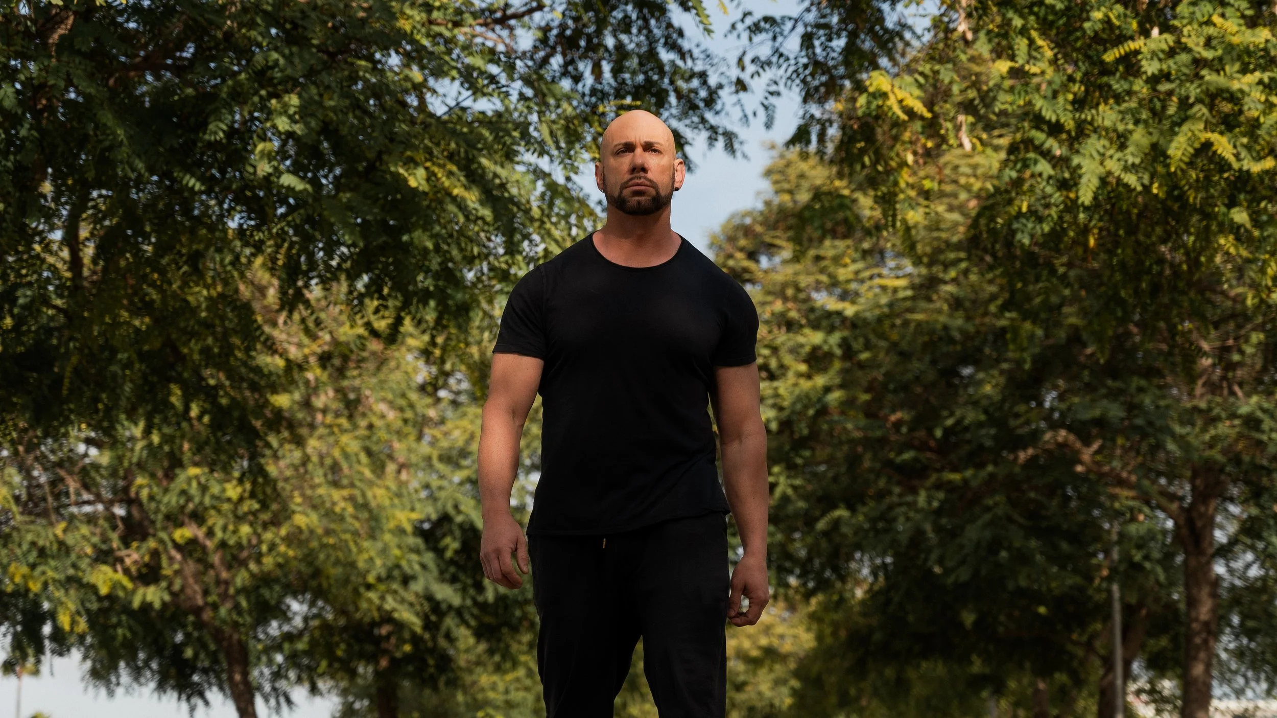 A muscular man with a shaved head and beard standing outdoors among green trees, wearing a black t-shirt and black pants.