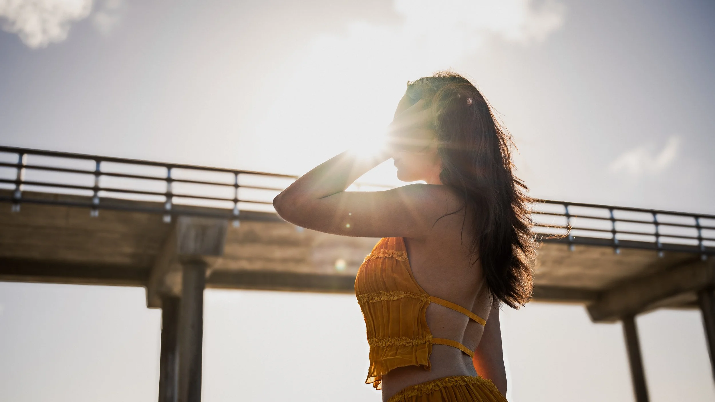 Woman standing outdoors in front of a bridge during sunset, wearing a yellow dress with ruffles and with her hand shielding her eyes from the sunlight.