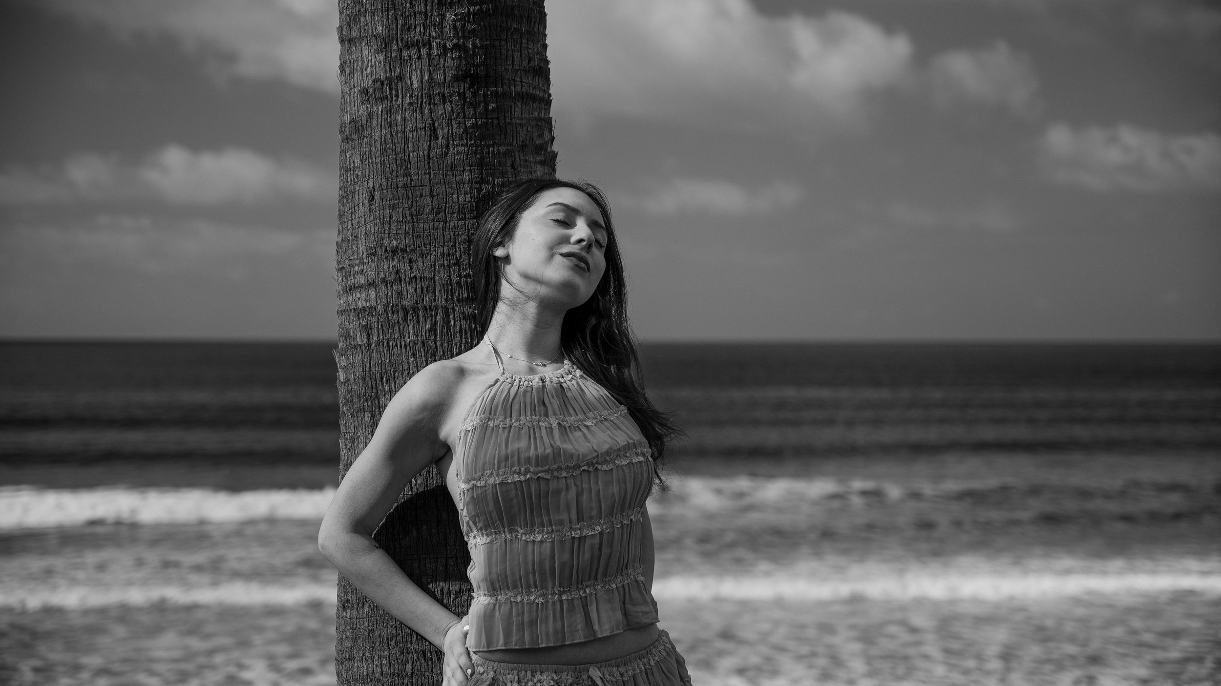 A woman in a sleeveless dress leaning against a palm tree on a beach, with the ocean and cloudy sky in the background, in black and white.