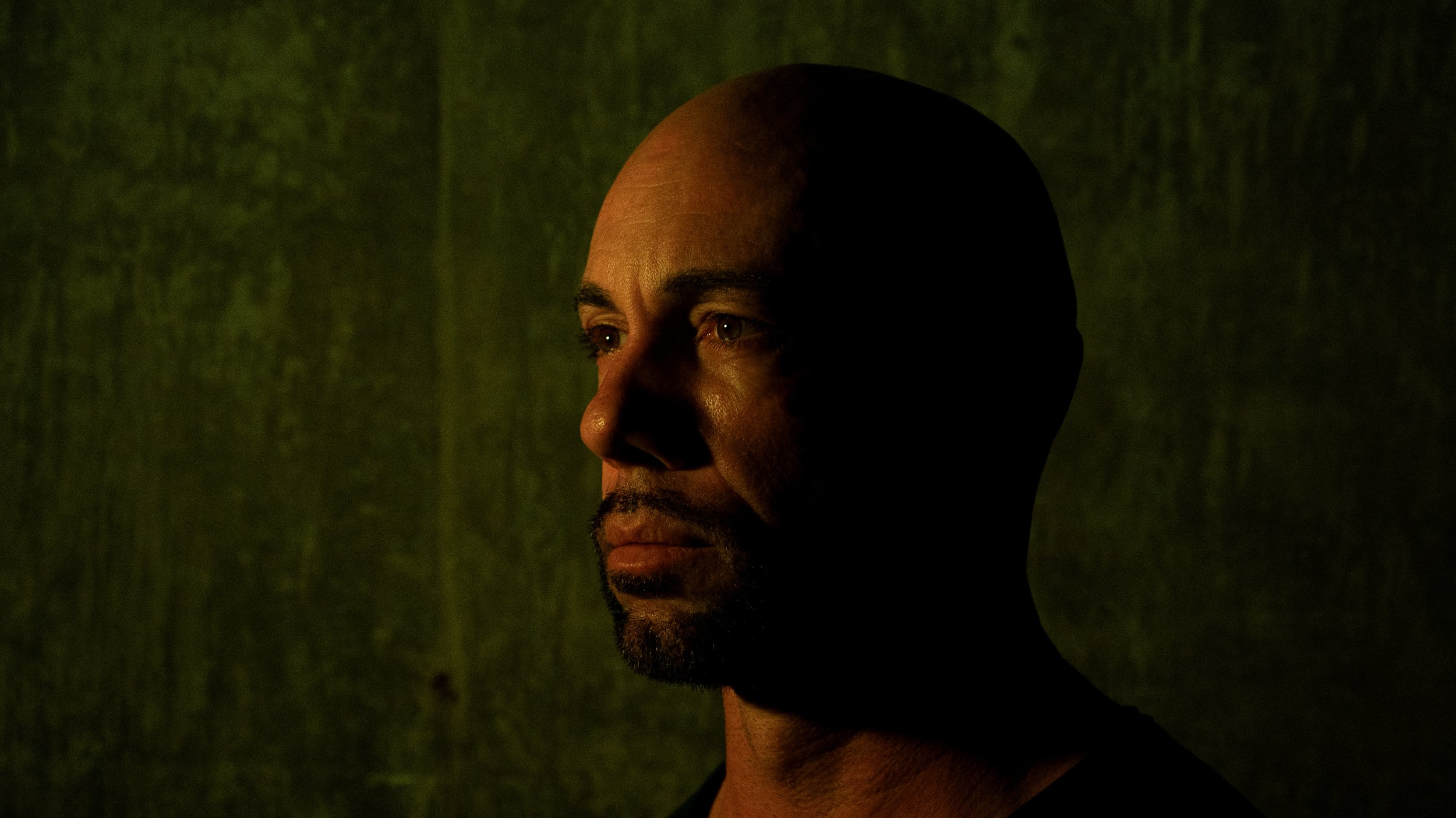 A man with a bald head and goatee looking to the left, illuminated from the front with a shadowed background