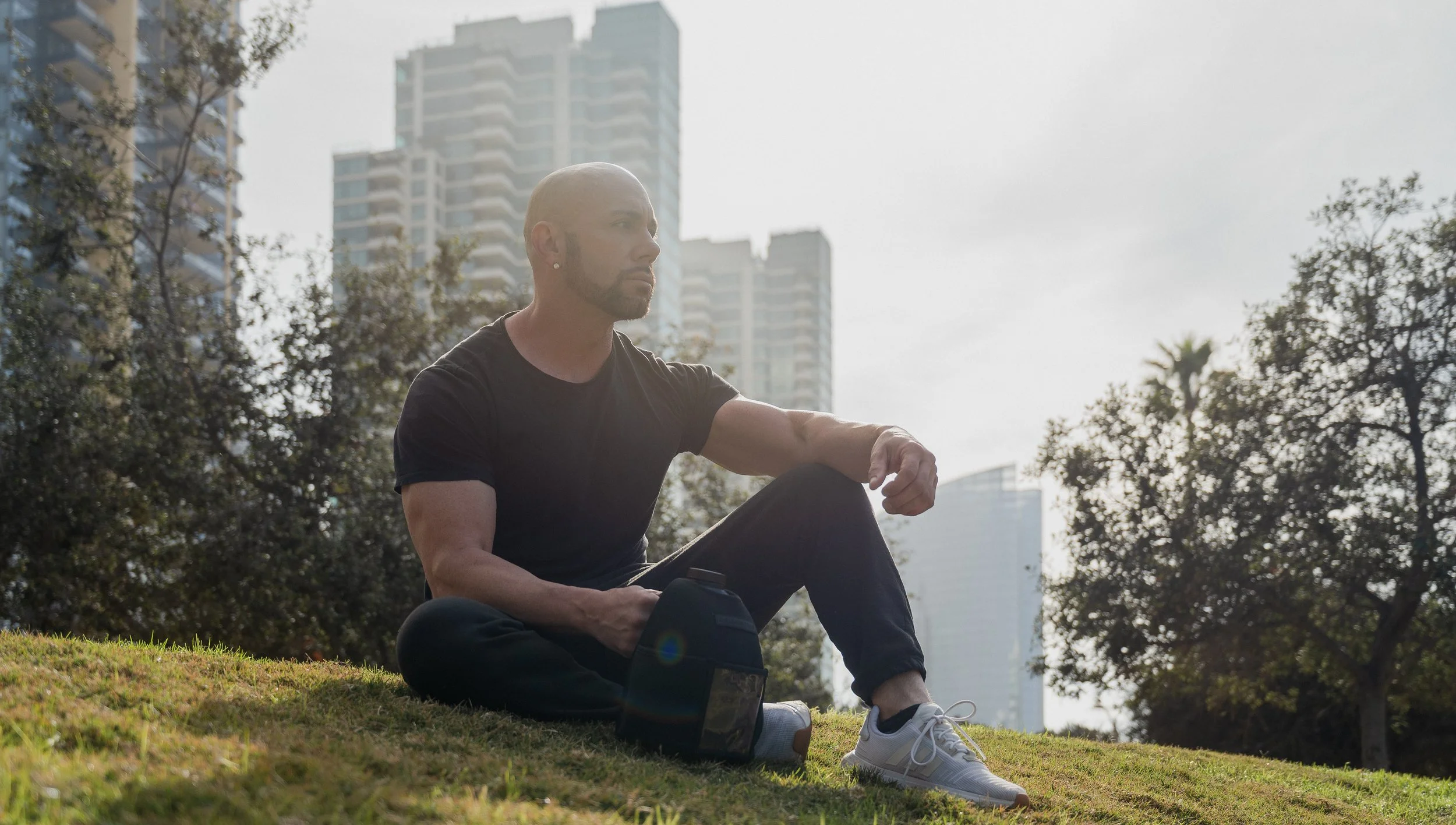 A person with a shaved head and beard sitting on grass in a park with city buildings in the background, holding a water bottle.