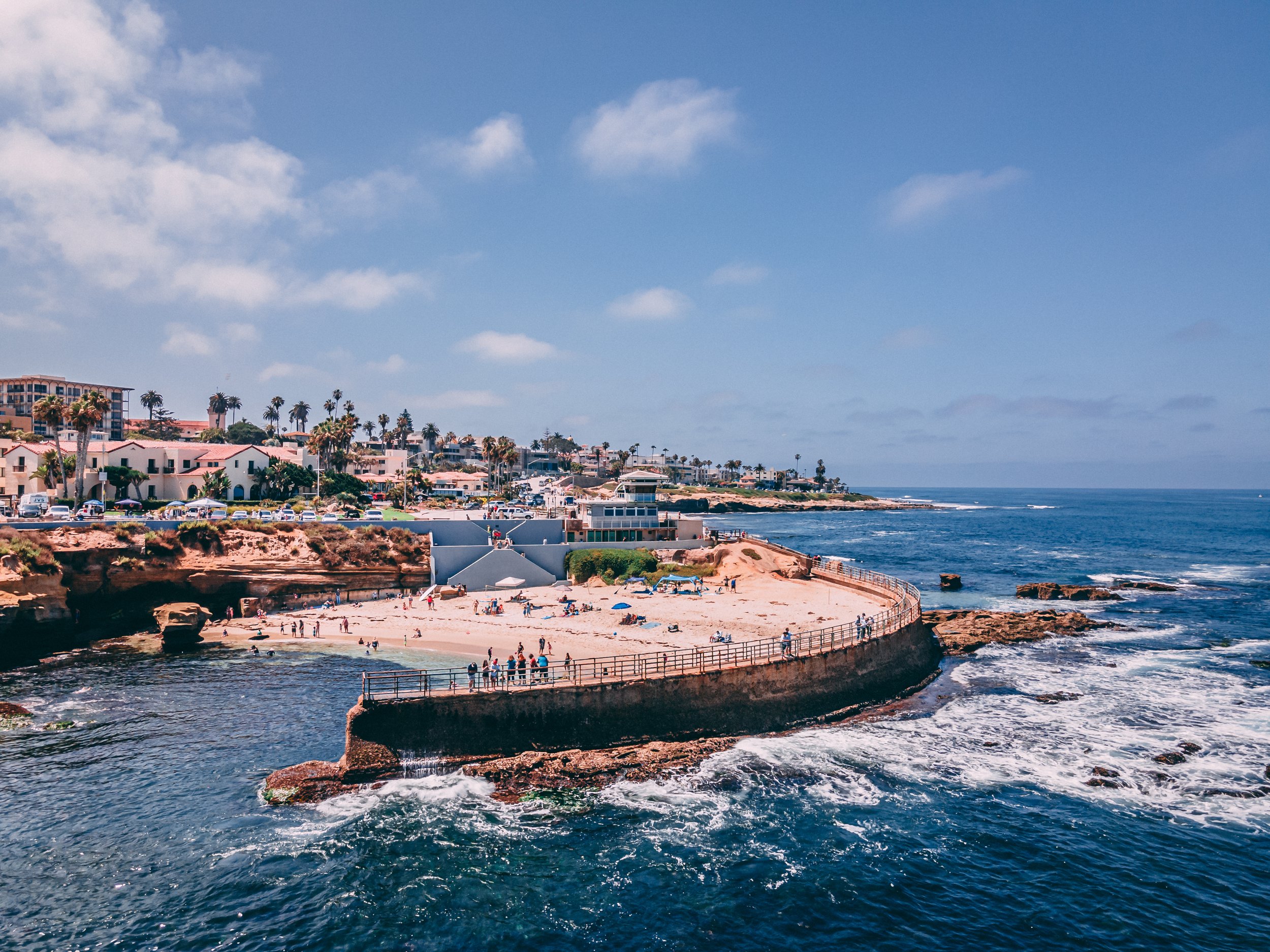 Coastal scene with sandy beach, ocean, and houses with palm trees under a partly cloudy sky.