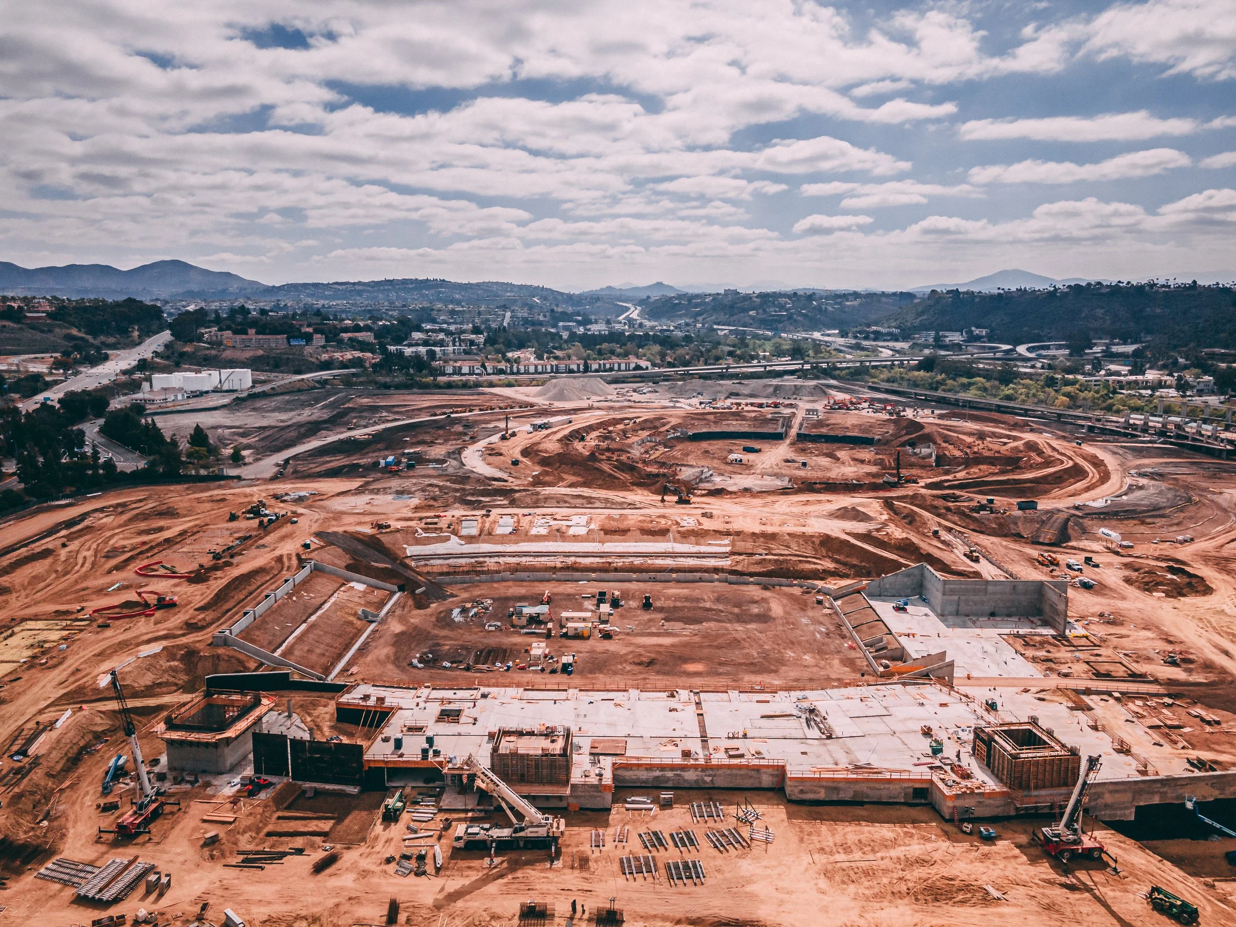 A large construction site at Snapdragon Stadium with building foundations, construction equipment, and dirt roads, surrounded by distant hills and a partly cloudy sky.