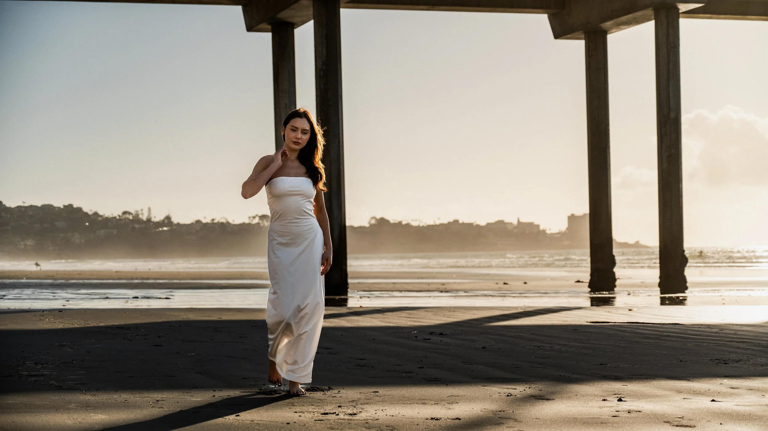 Woman in a white dress walking barefoot on the beach near a pier at sunset.