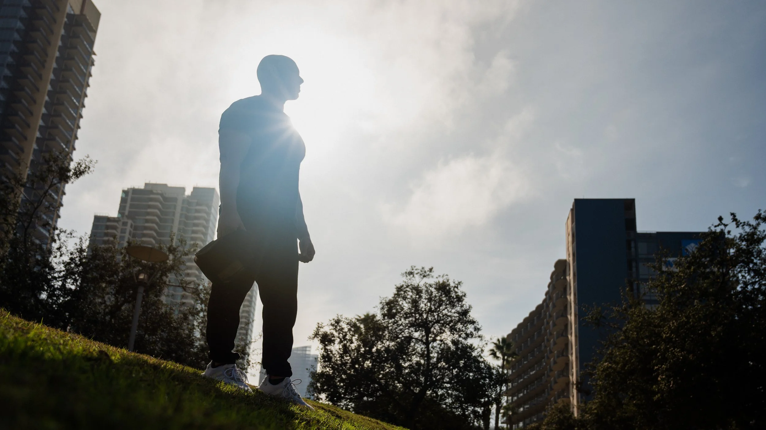 Silhouette of a man walking in an urban park during daytime, with tall buildings and trees in the background, and the sun shining behind him.