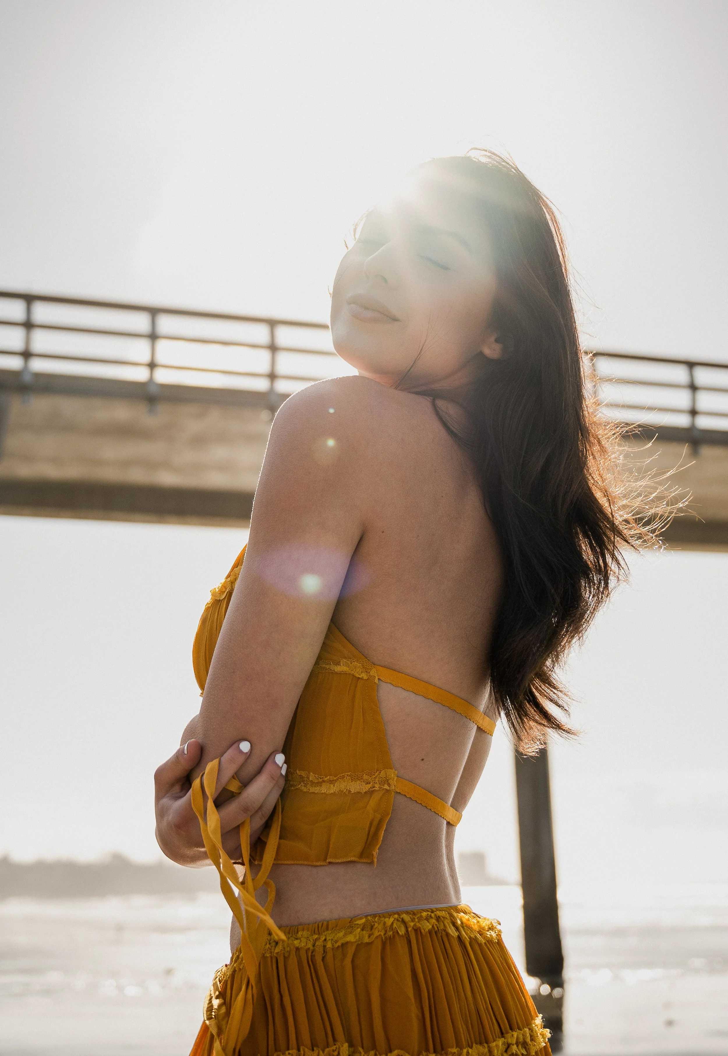 A woman in a yellow dress, standing outdoors near water, with the sun shining behind her, creating a lens flare and a bright glow around her head.
